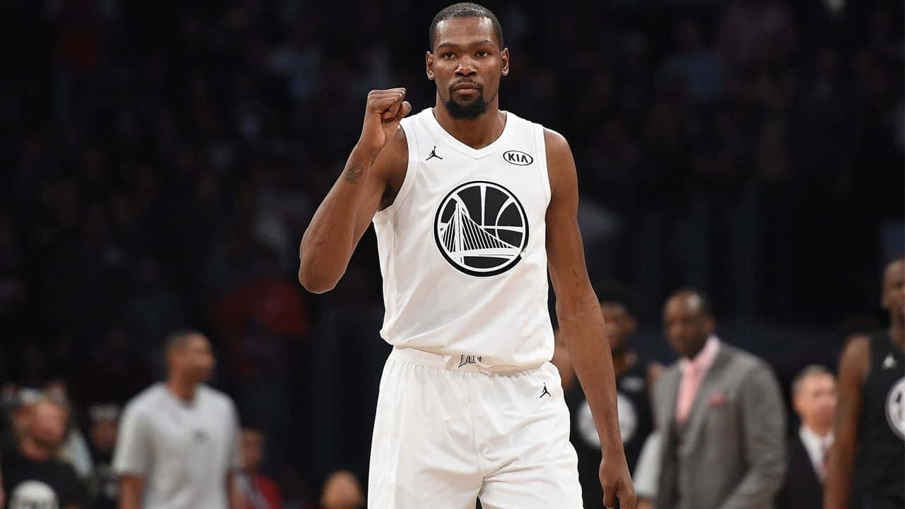 凱文·杜蘭特 桌布 壁紙 — 籃球運動員Kevin Durant身穿白色勇士隊球衣，在明亮的燈光下於球場內舉起拳頭。