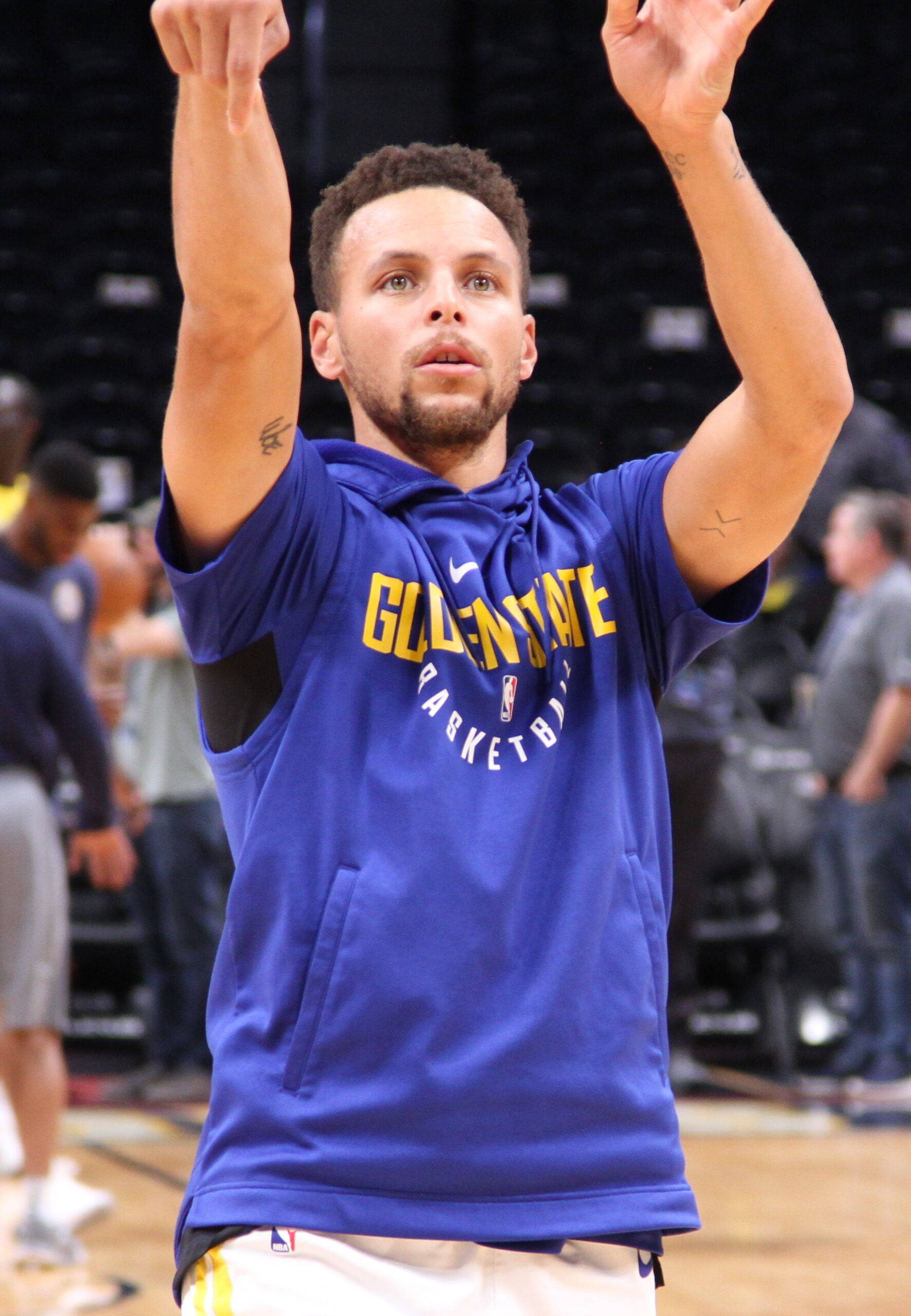 NBA 桌布 壁紙 — 籃球運動員史蒂芬柯瑞身穿藍色金州勇士連帽上衣，在硬木球場上專注熱身，高舉雙臂。