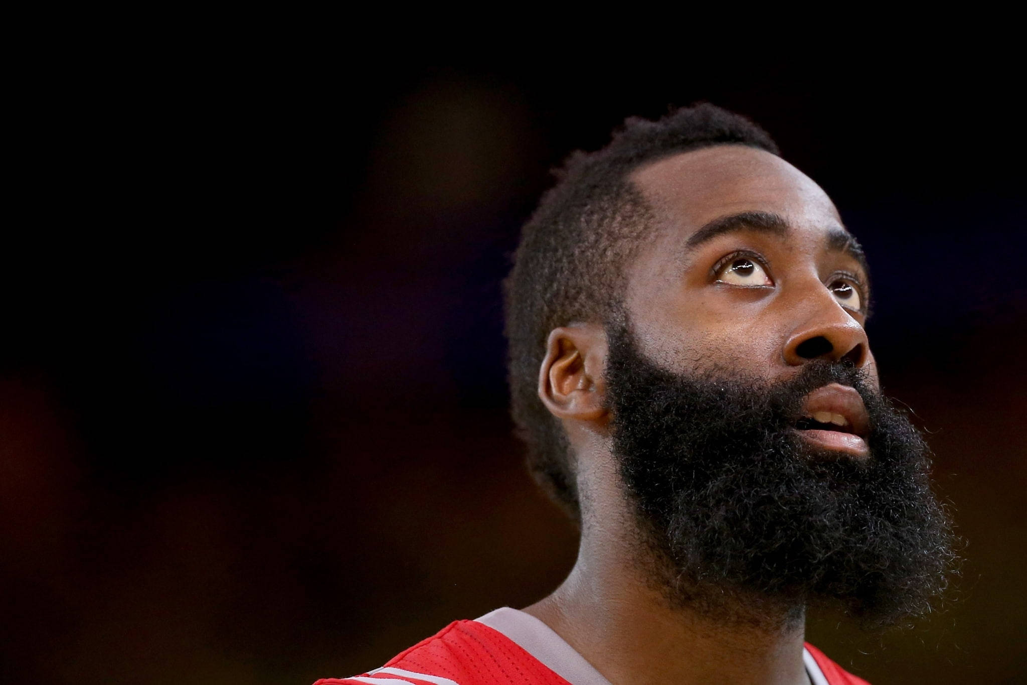 James Harden 桌布 壁紙 — 籃球運動員詹姆斯哈登身穿紅色球衣的特寫肖像，在黑暗模糊的背景前向上凝視，神情專注。