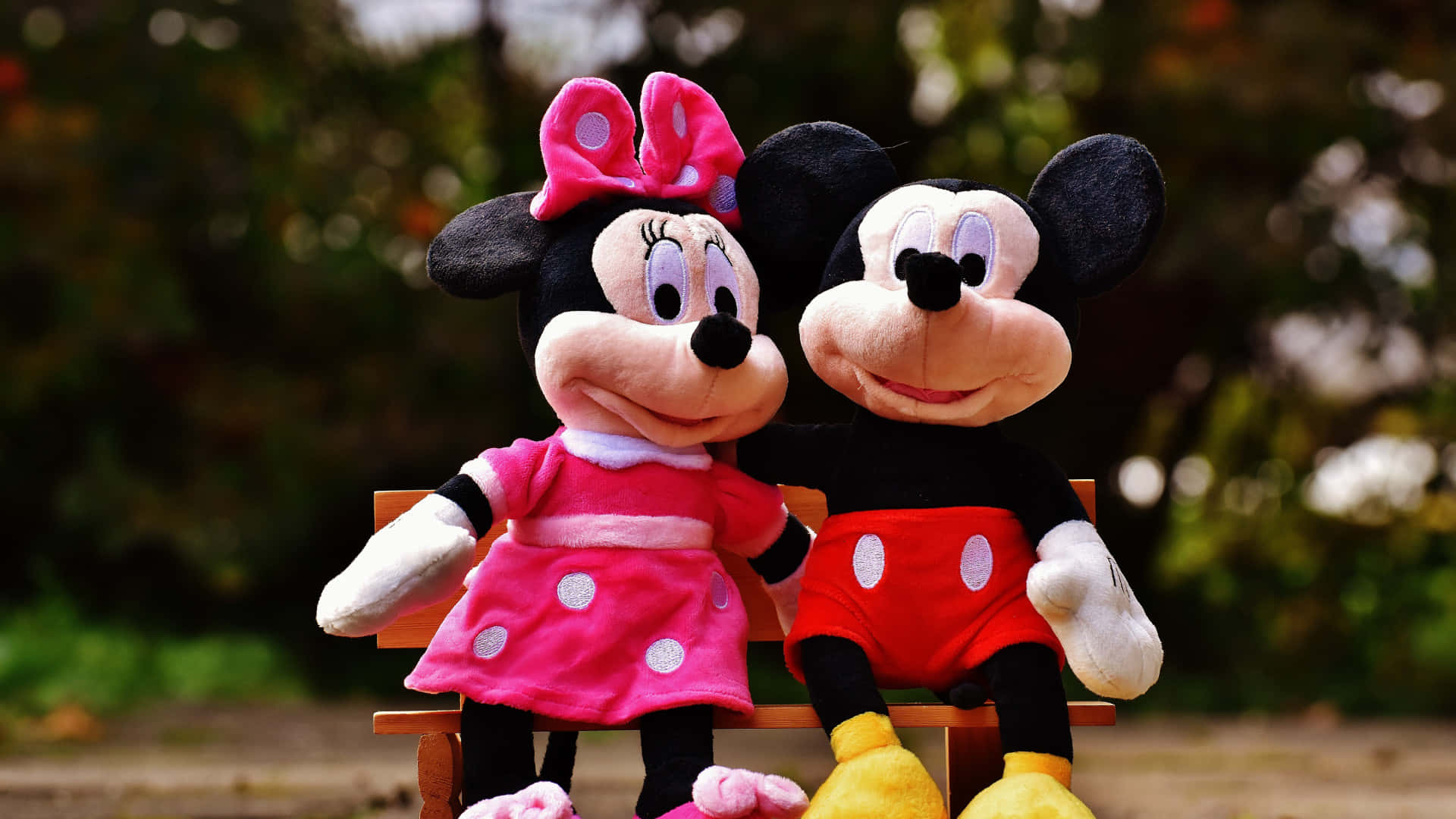 Mickey And Minnie Mouse 桌布 壁紙 — 米奇和米妮老鼠毛絨玩具一起坐在陽光普照的公園木長椅上，身後是模糊的綠色樹葉。