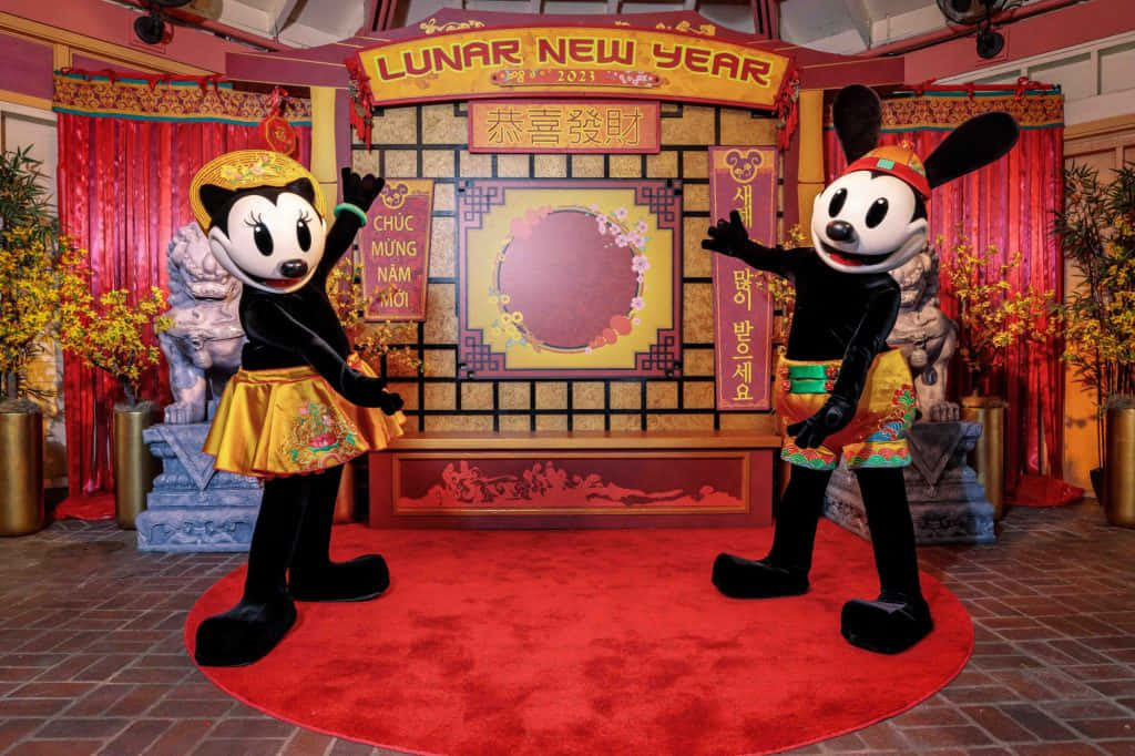 桌布 壁紙 — 米奇和米妮老鼠穿著喜慶的農曆新年服裝，在裝飾著紅色窗簾和獅子雕像的舞台前愉快地擺姿勢。