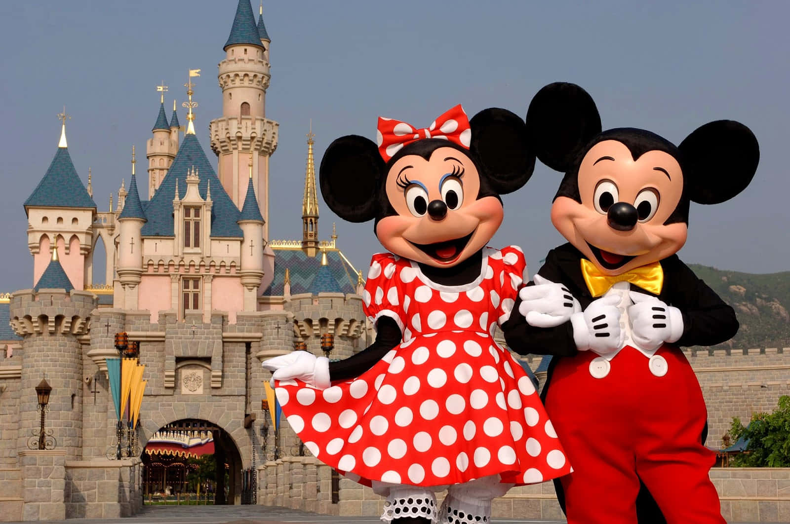 Mickey And Minnie Mouse 桌布 壁紙 — 米奇和米妮老鼠穿著鮮豔的服裝，在晴朗的天空下，歡快地站在宏偉的童話城堡前。