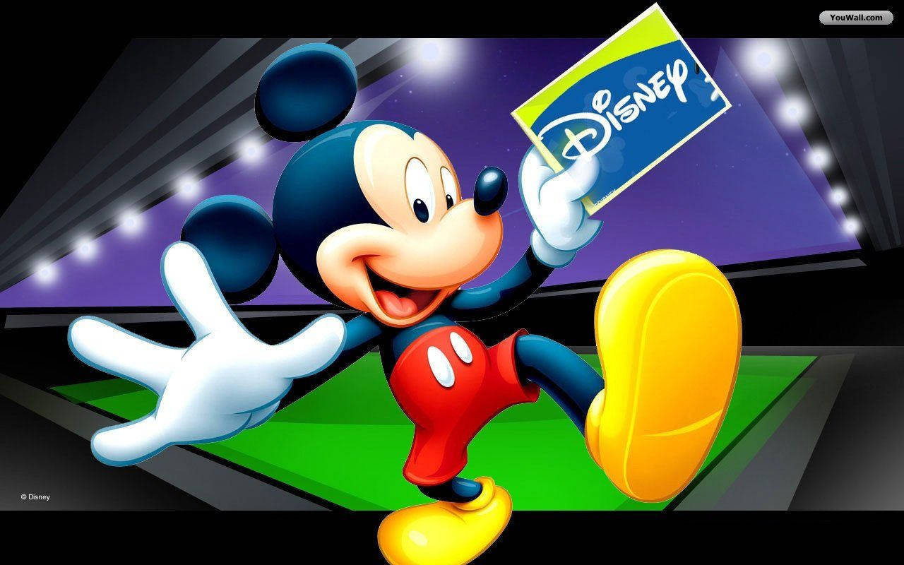 Mickey 桌布 壁紙 — 米奇老鼠愉快地揮手，並在綠色地板上明亮的舞台燈光下拿著迪士尼標誌。