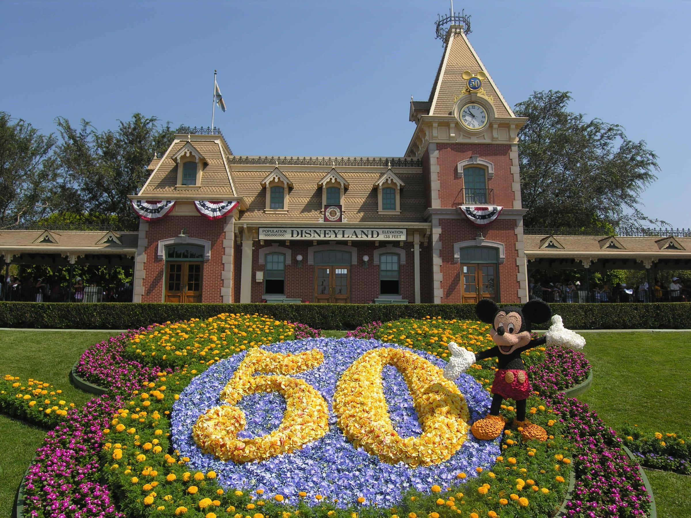 Disneyland 桌布 壁紙 — 米奇老鼠站在迪士尼樂園標誌性火車站前，旁邊是拼出「50」字樣的鮮豔花卉佈置，背景是晴朗的藍天。