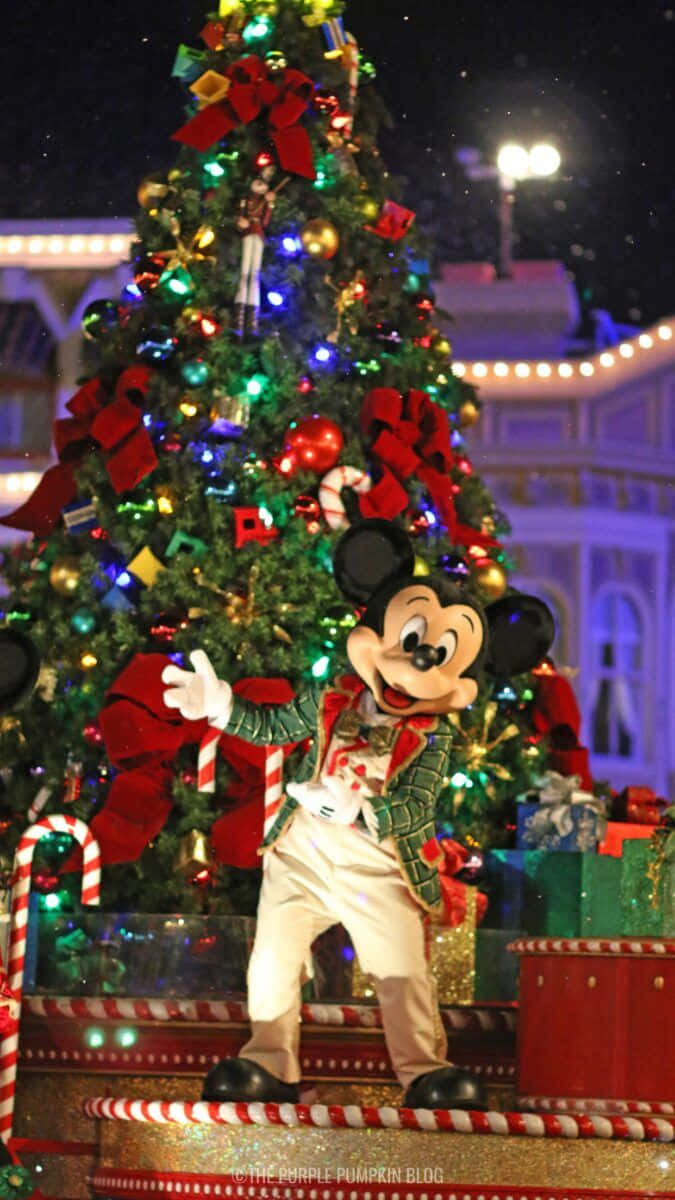 Disney Christmas iPad 桌布 壁紙 — 米奇老鼠身穿節日服裝，在夜晚於一棵裝飾著紅色蝴蝶結和拐杖糖裝飾、燈火通明的聖誕樹旁愉快地揮手。