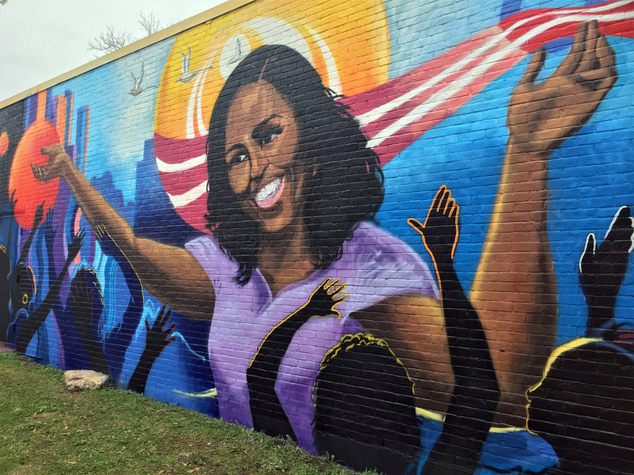 Michelle 桌布 壁紙 — 米歇爾·奧巴馬微笑揮手的鮮豔壁畫，背景是色彩繽紛的城市景觀，有舉起的手和愛國元素。