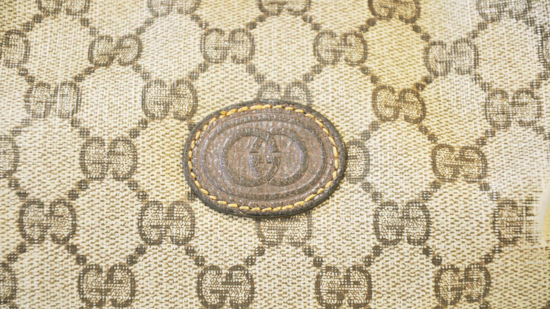 Gucci 桌布 壁紙 — 米色和棕色Gucci字母組合帆布近距離特寫，中央有一個橢圓形皮革標誌貼片。