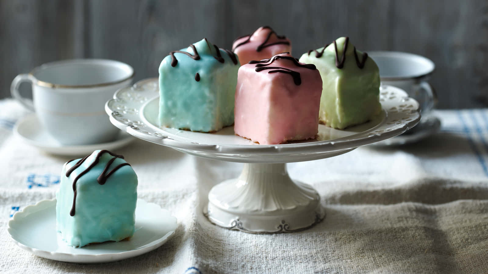 Cake Fondant 桌布 壁紙 — 粉彩方形蛋糕配上巧克力淋醬，優雅地展示在白色底座架上，旁邊放著茶杯，置於有紋理的亞麻桌布上。