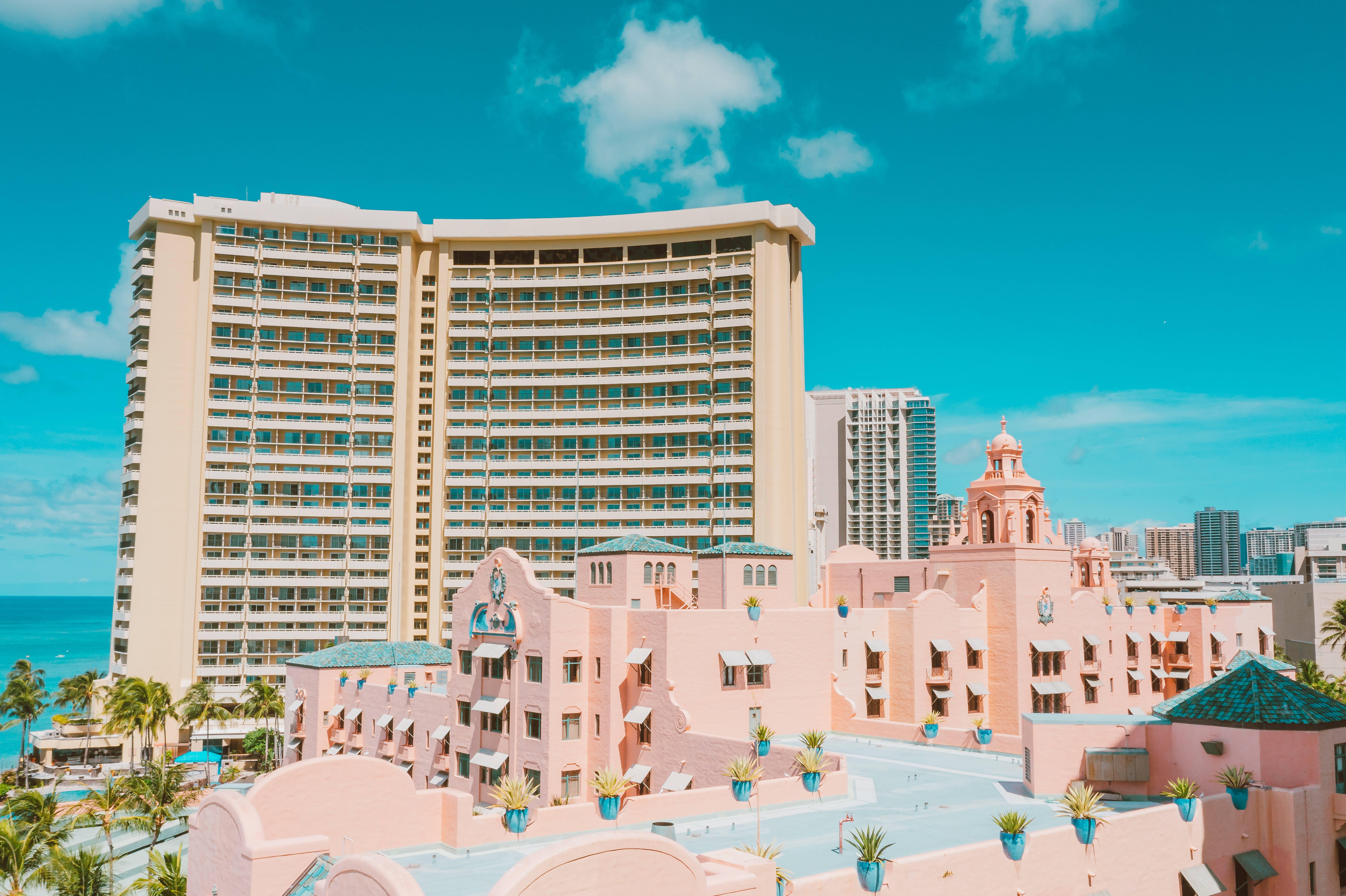 Cute Pastel Aesthetic 桌布 壁紙 — 粉彩粉紅色度假村建築，配以綠松石色屋頂和棕櫚樹，排列在明亮的綠松石色海洋旁，上方是帶有蓬鬆雲朵的鮮豔藍天。