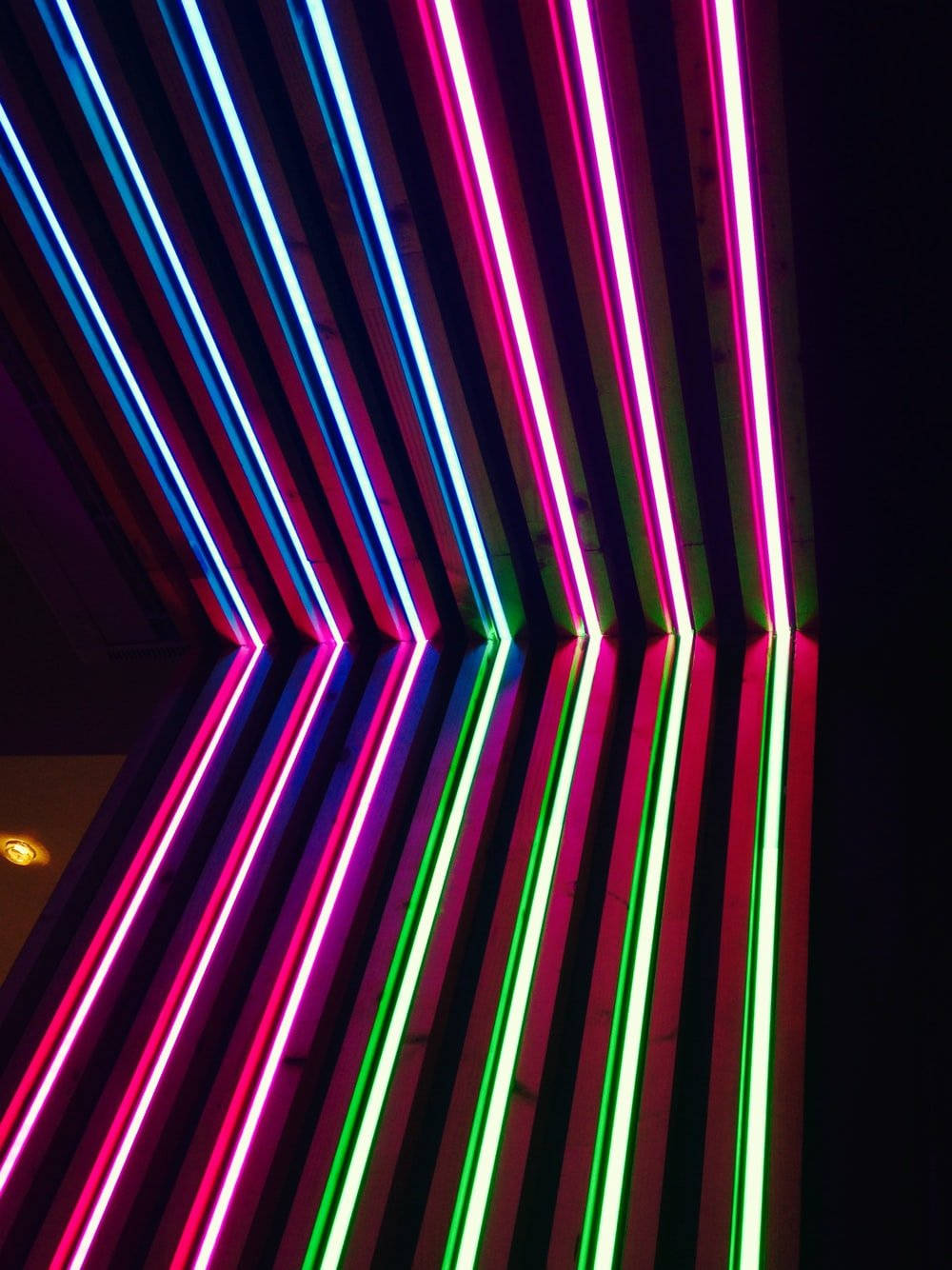 Neon Light 桌布 壁紙 — 粉紅、藍色和綠色的鮮豔霓虹管在黑暗的天花板上對角線延伸，創造出未來感的發光圖案。