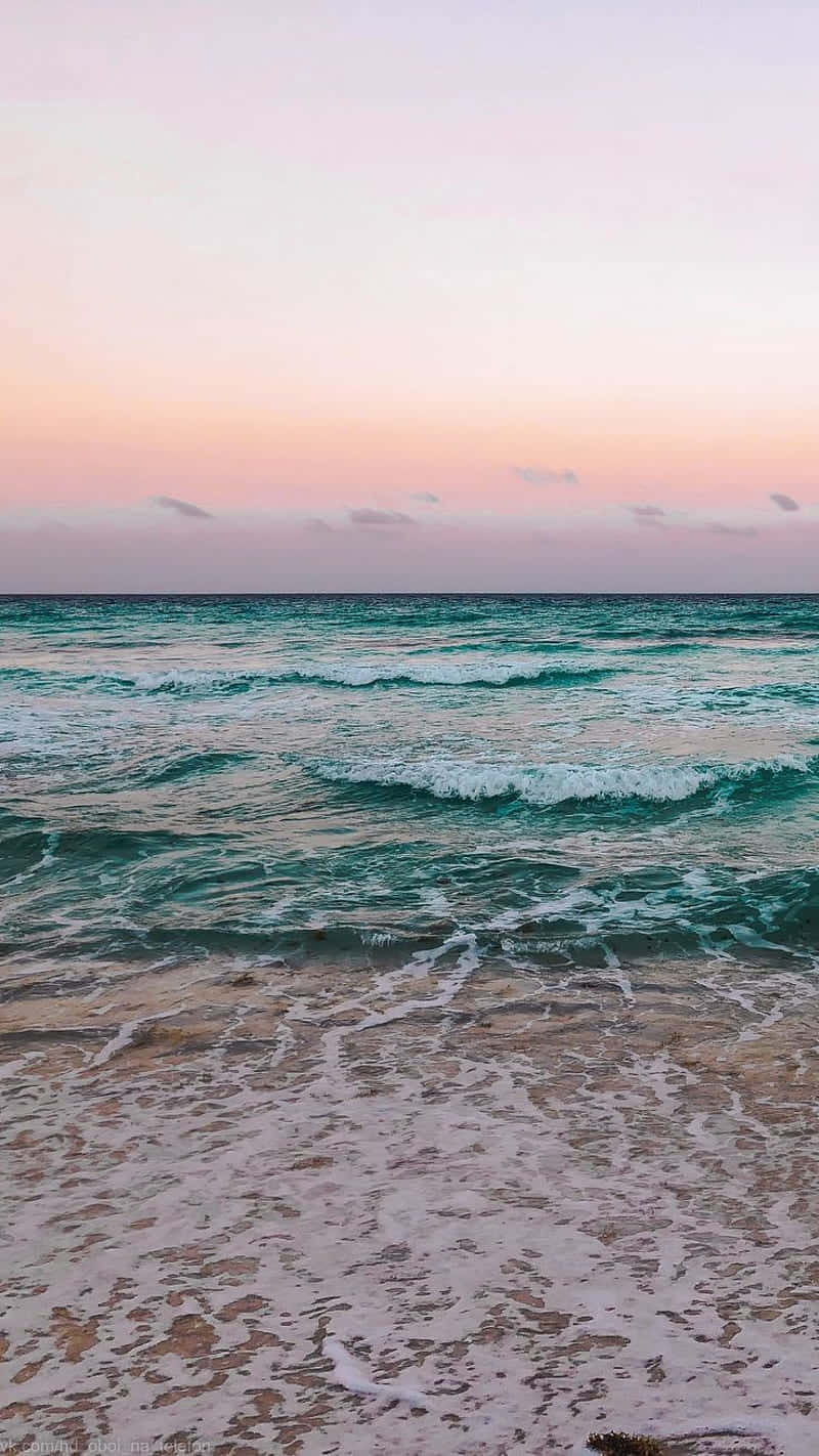 Beach Aesthetic 桌布 壁紙 — 粉紅和蜜桃色的日落色調融合在碧綠的海浪上，海浪拍打著沙灘。