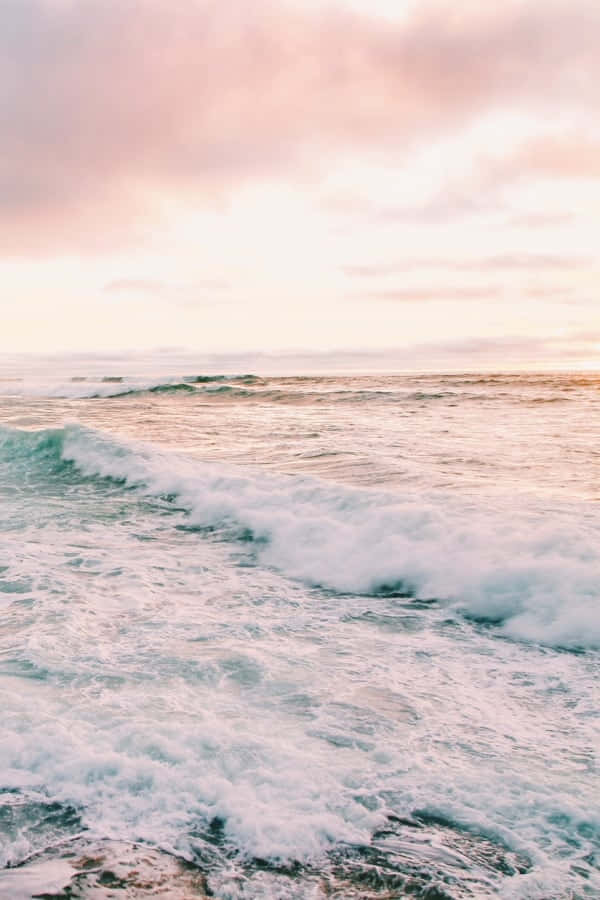 Beach Aesthetic 桌布 壁紙 — 粉紅和蜜桃色的雲彩在翻騰的海洋上發光，白色的泡沫波浪向岸邊滾動。