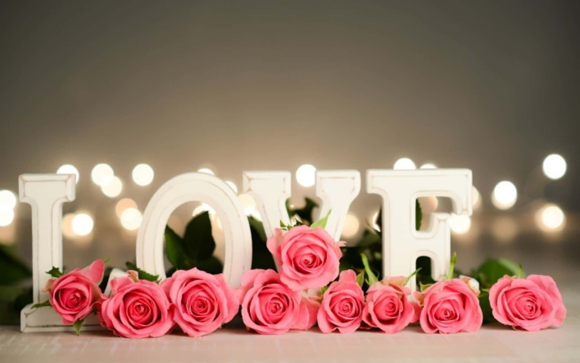 愛心 桌布 壁紙 — 粉紅玫瑰排列在白色 'LOVE' 字母前，背景有溫暖的散景燈光。