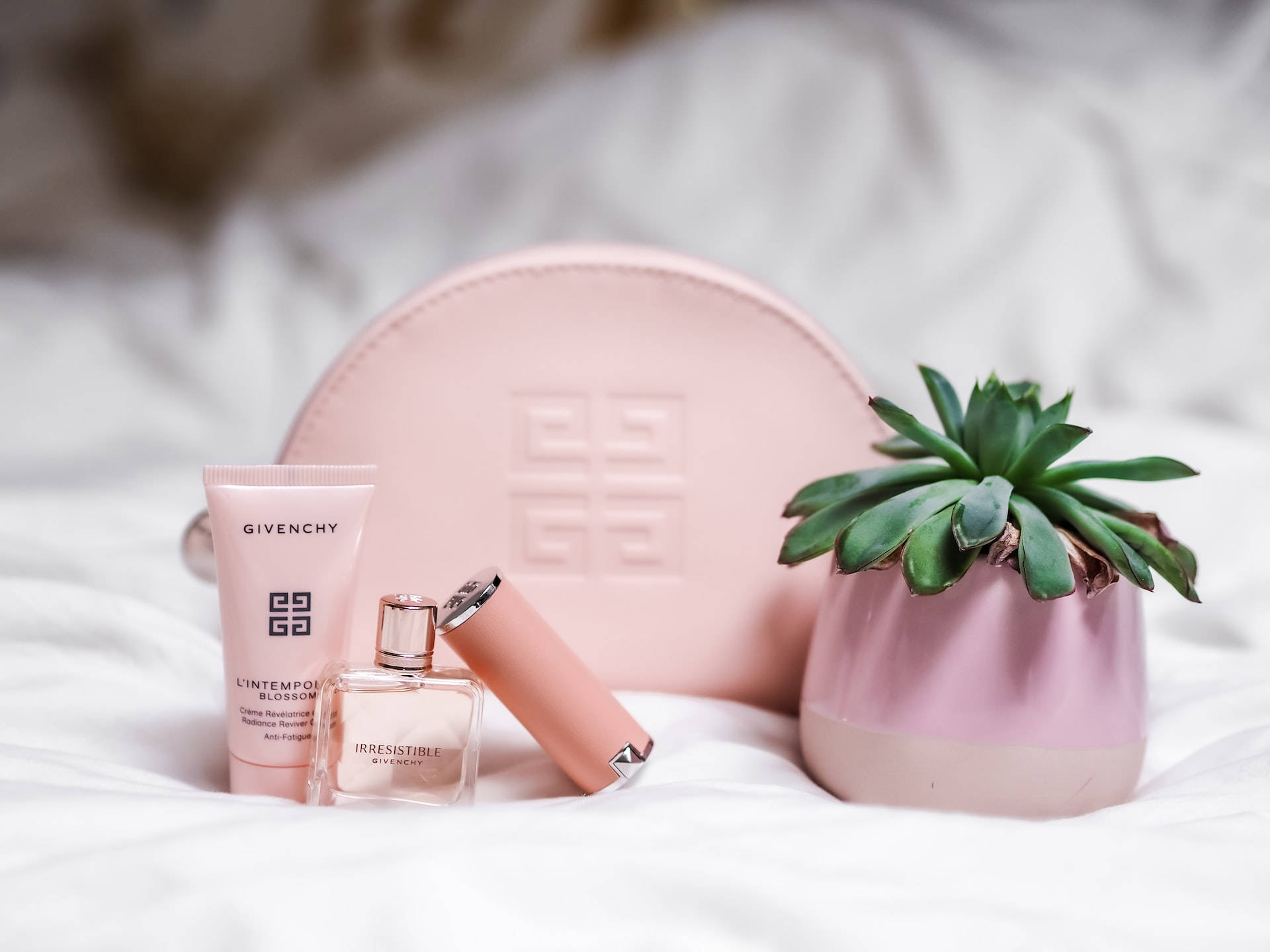 玫瑰金美學 桌布 壁紙 — 粉紅色 Givenchy 美容產品和一個多肉植物放在相配的花盆中，置於柔軟的白色床單上。