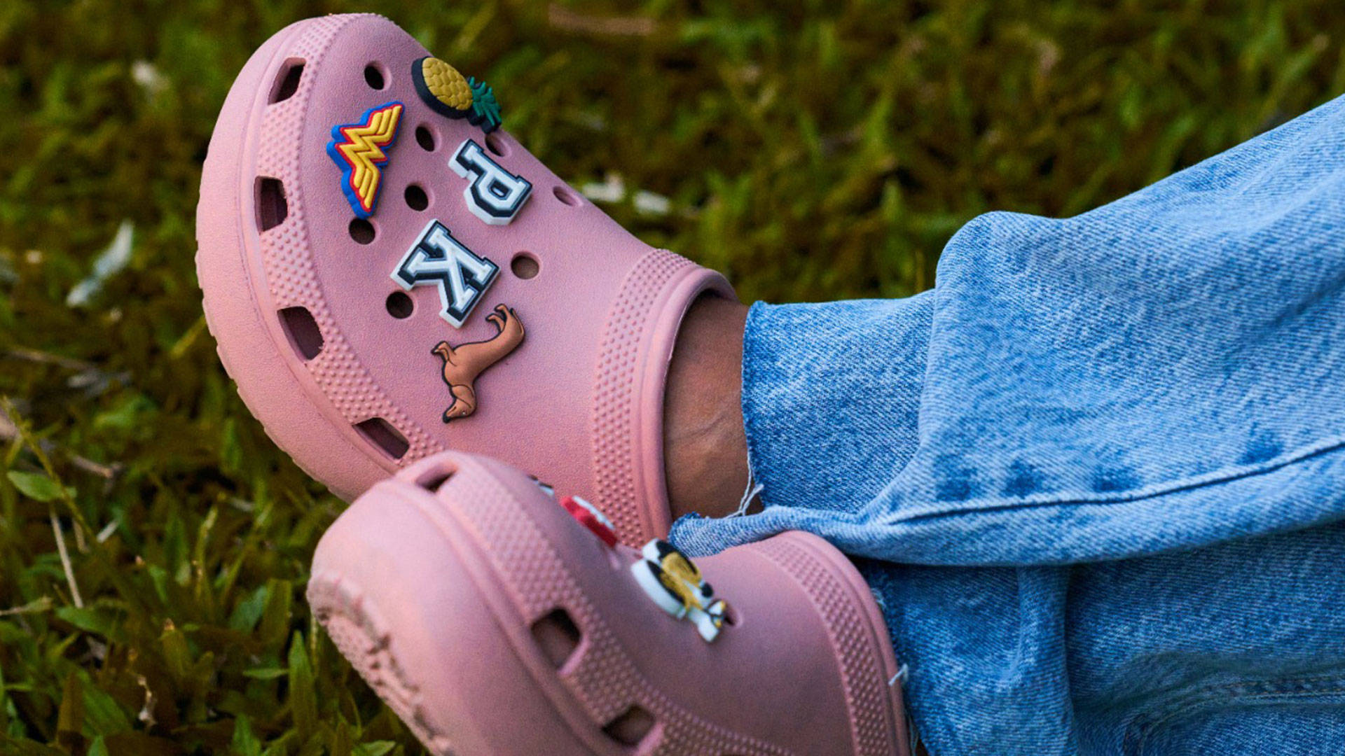 Crocs 桌布 壁紙 — 粉紅色Crocs近距離特寫，飾有彩色補丁，放在綠色苔蘚上，旁邊是藍色牛仔褲。