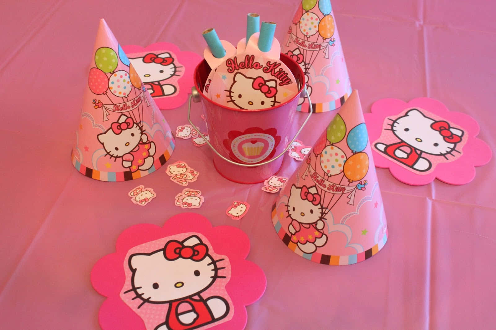 Hello Kitty Party 桌布 壁紙 — 粉紅色Hello Kitty派對用品，包括帽子、吸管和花朵形狀裝飾，排列在粉紅色桌布上。