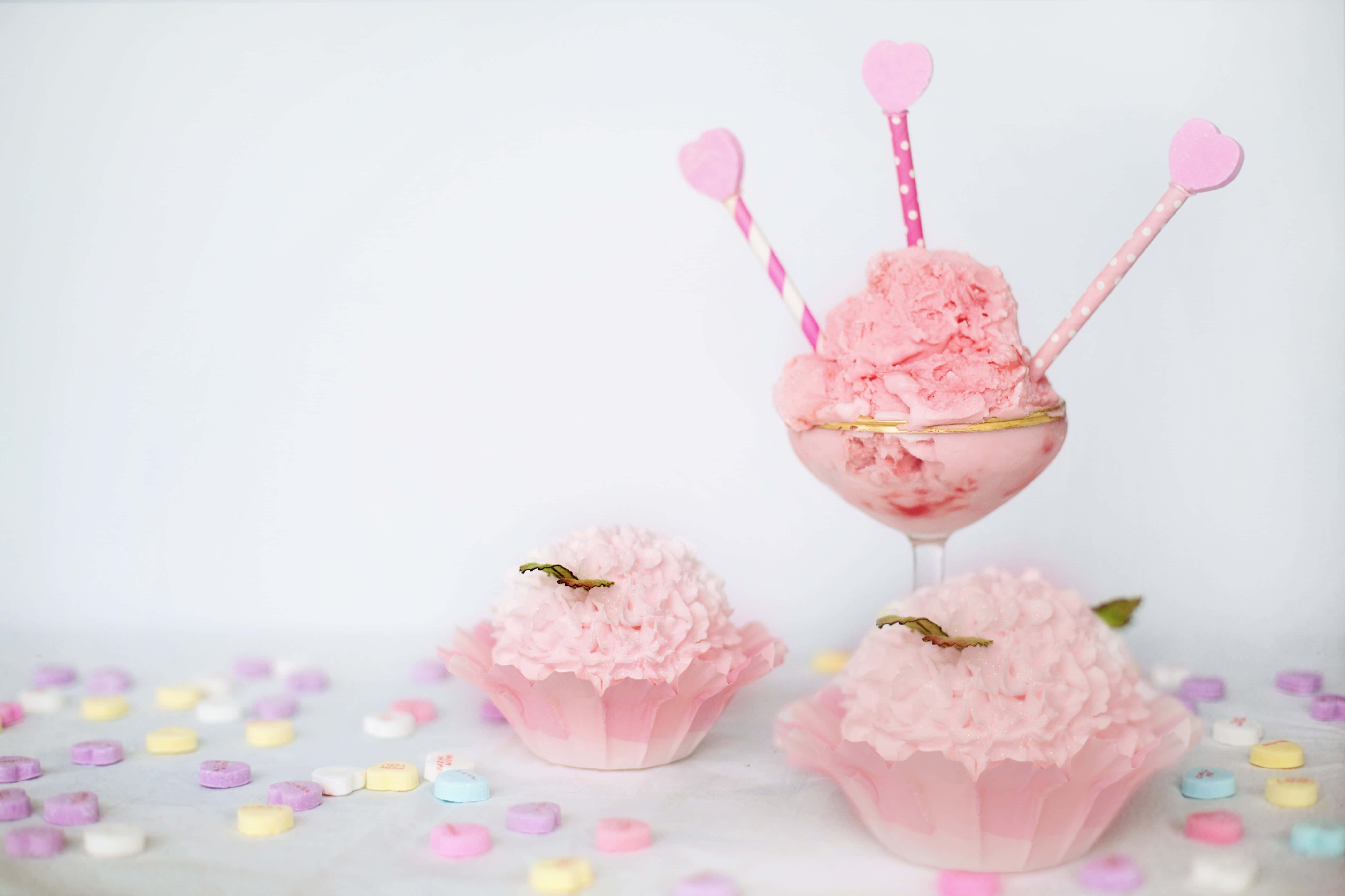 Pink Cupcakes 桌布 壁紙 — 粉紅色冰淇淋放在玻璃杯裡，配上心形吸管和杯子蛋糕，周圍散落著色彩繽紛的心形糖果，置於白色表面上。