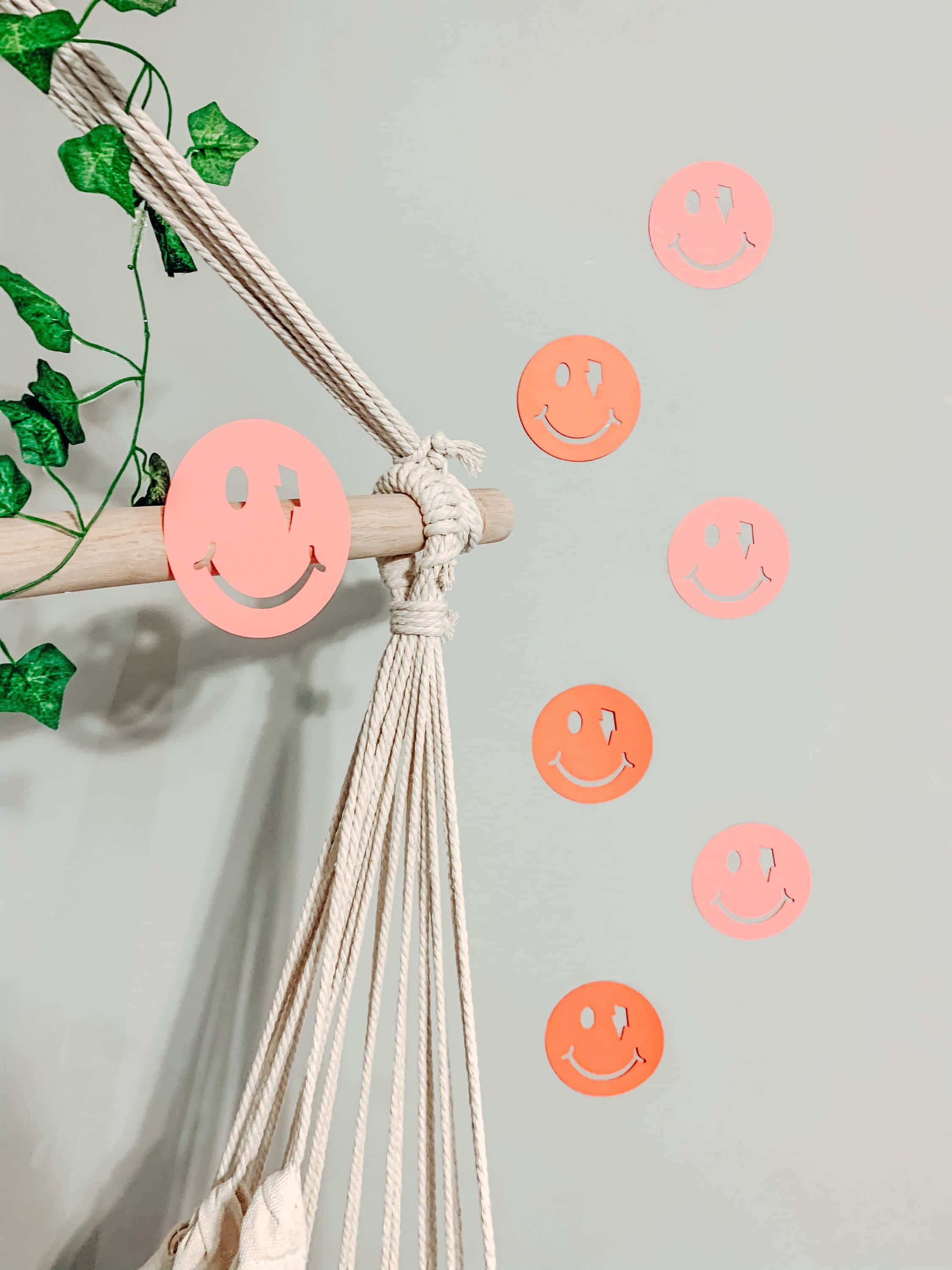 Aesthetic Smiley Face 桌布 壁紙 — 粉紅色和橙色的紙製笑臉在柔和的灰色牆壁旁邊漂浮，旁邊掛著一個帶有綠色常春藤的流蘇掛飾。