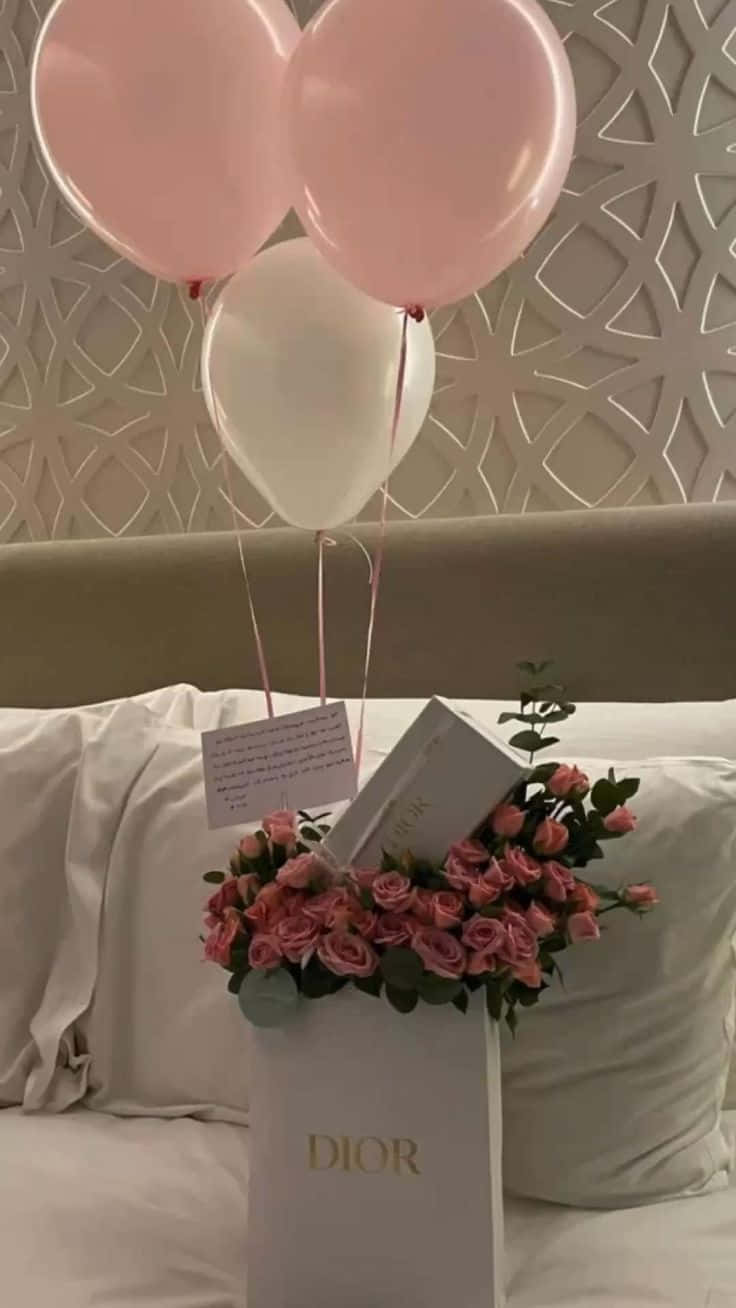 Birthday Message 桌布 壁紙 — 粉紅色和白色的氣球飄浮在一個裝滿粉紅色玫瑰的Dior禮物袋上方，放在一張背後有圖案牆壁的床上。