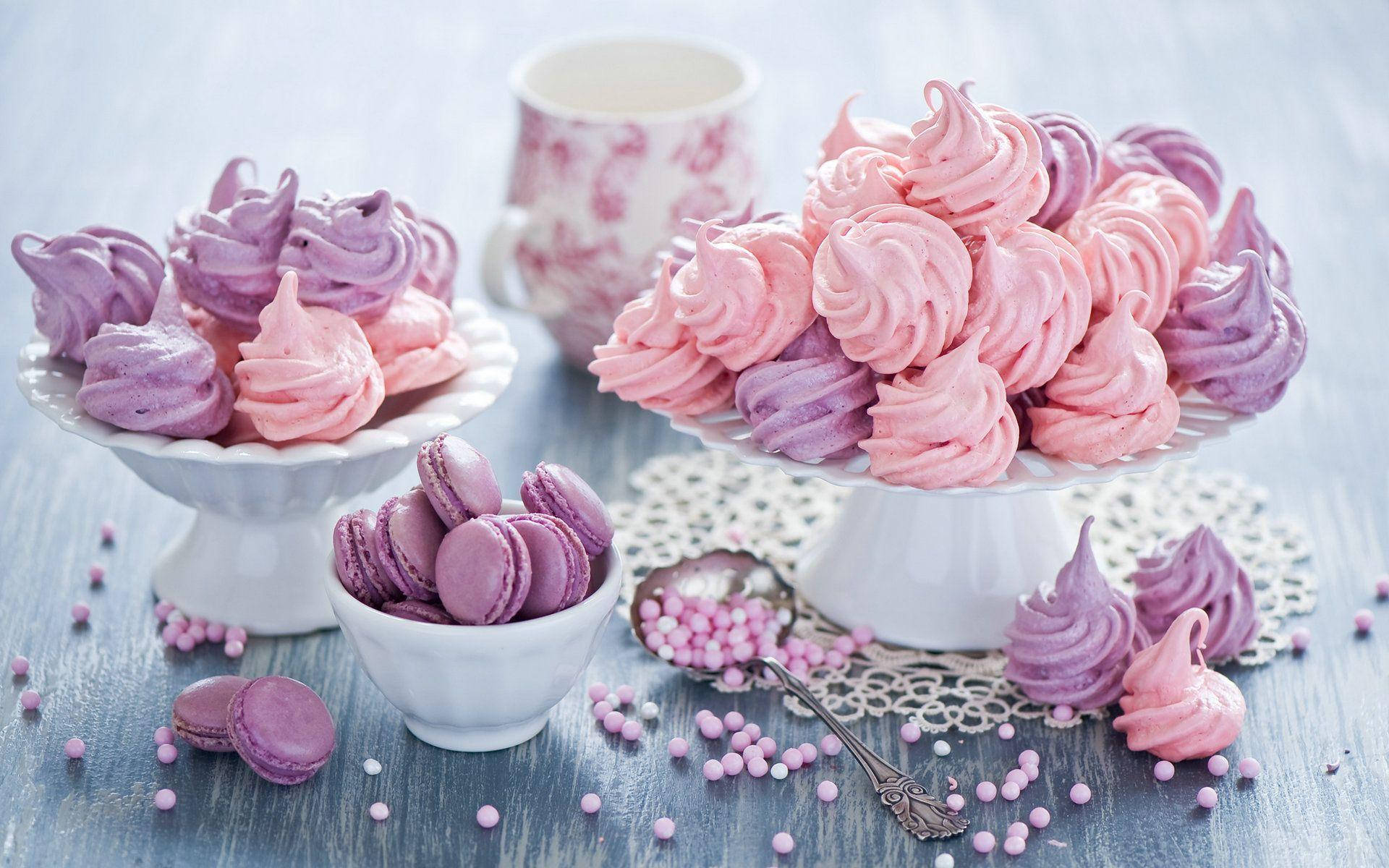 Desserts 桌布 壁紙 — 粉紅色和紫色的馬卡龍和蛋白霜放在白色架子上，撒上糖珠，放在質樸的藍色木桌上。