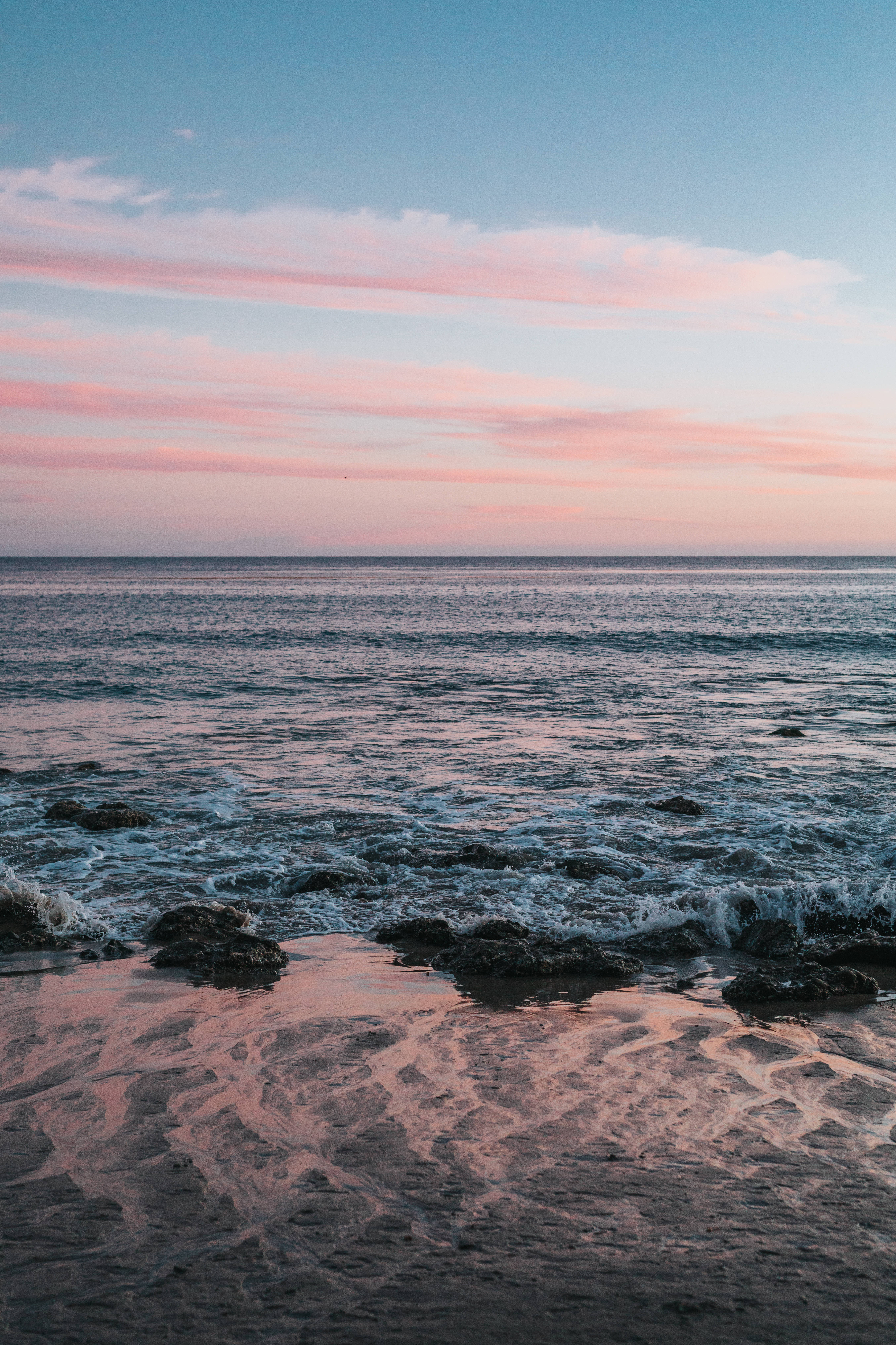 malibu iPhone 桌布 壁紙 — 粉紅色和藍色的日落色調塗滿天空，並在海浪拍打濕岩石和沙灘時泛起漣漪。