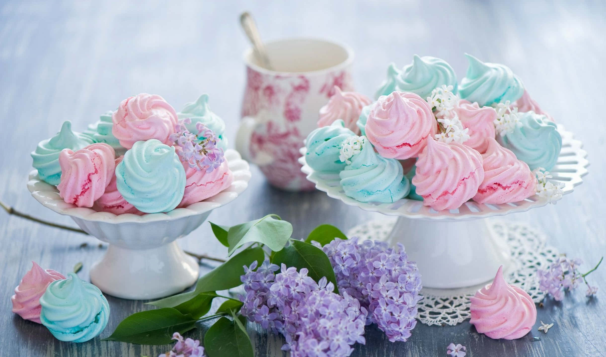 2440x1440 Desserts 桌布 壁紙 — 粉紅色和藍色蛋白甜餅放在白色架子上，旁邊有紫色丁香花和一個花卉圖案的杯子，擺在質樸的木桌上。