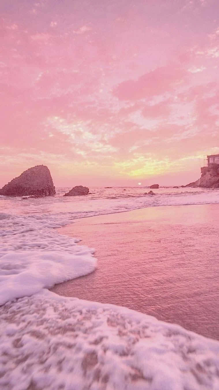 Pink Beach 桌布 壁紙 — 粉紅色日落灑落在寧靜的海灘上，泡沫般的海浪拍打著海岸，遠處的岩石在柔和的天空下。