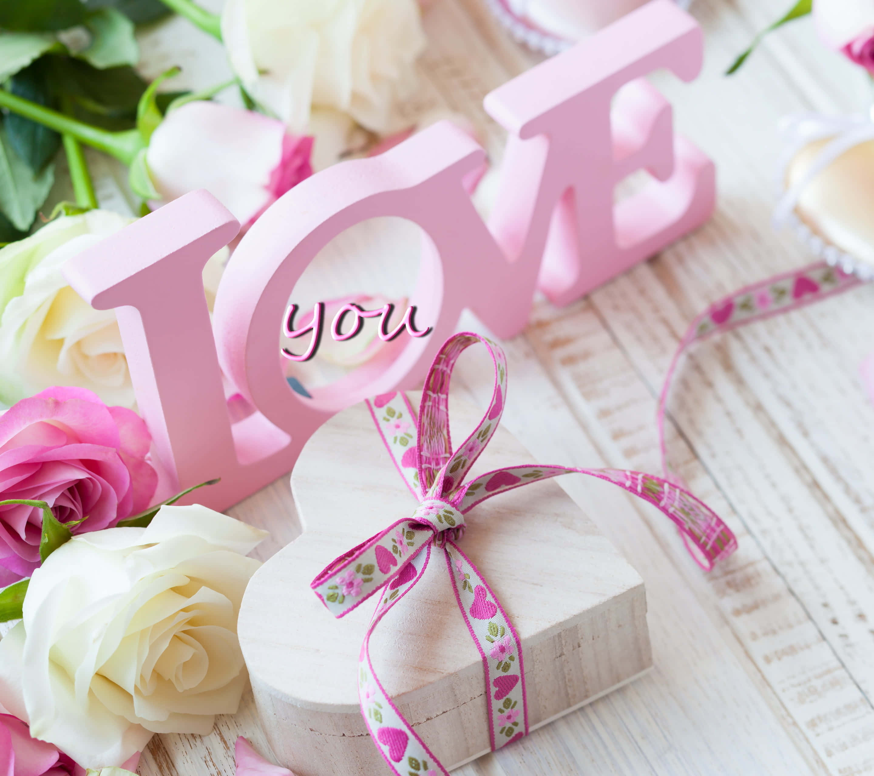 Pink Roses 桌布 壁紙 — 粉紅色木製字母拼出 LOVE，圍繞一個心形禮物盒，繫有圖案絲帶，周圍環繞著柔和的粉紅色和白色玫瑰，置於鄉村風木質表面上。