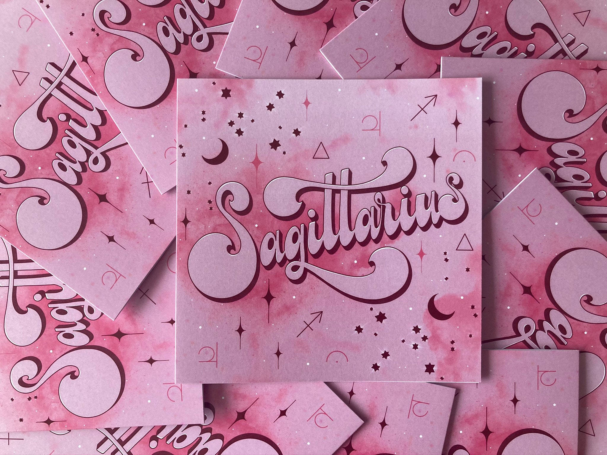 sagittarius 桌布 壁紙 — 粉紅色水彩卡片，帶有優雅的人馬座字體和宇宙符號，散落在柔軟夢幻的一堆中。
