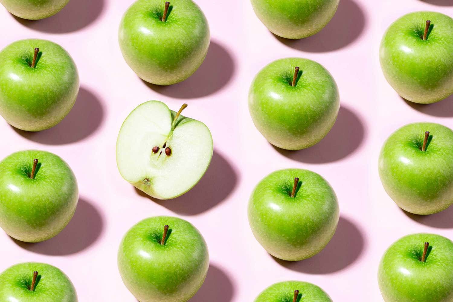 Fruits 桌布 壁紙 — 粉紅色背景上排列著充滿活力的青蘋果網格，其中一個切半的蘋果在中間露出種子。