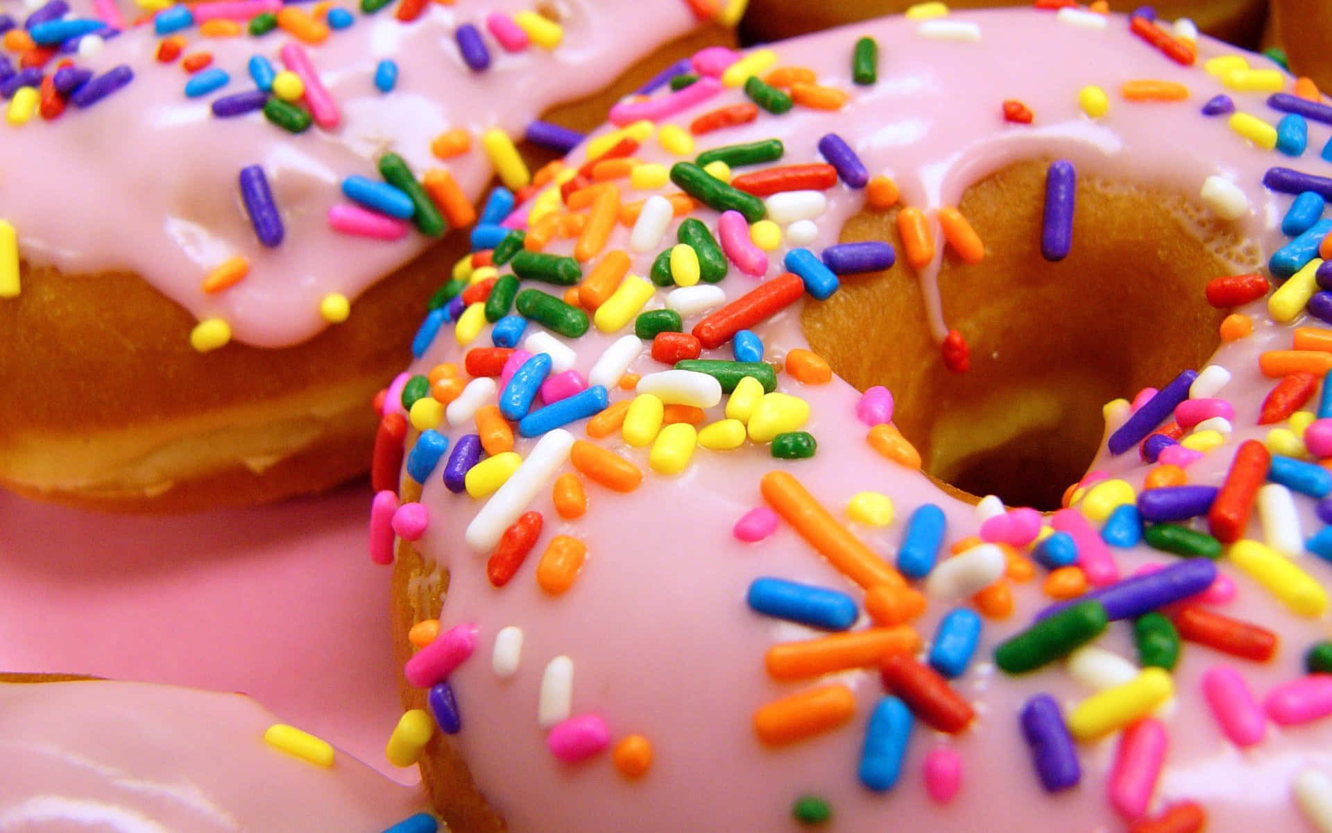 Cute Donut 桌布 壁紙 — 粉紅色表面上覆蓋著彩色彩虹糖屑的光澤粉紅色糖霜甜甜圈特寫。