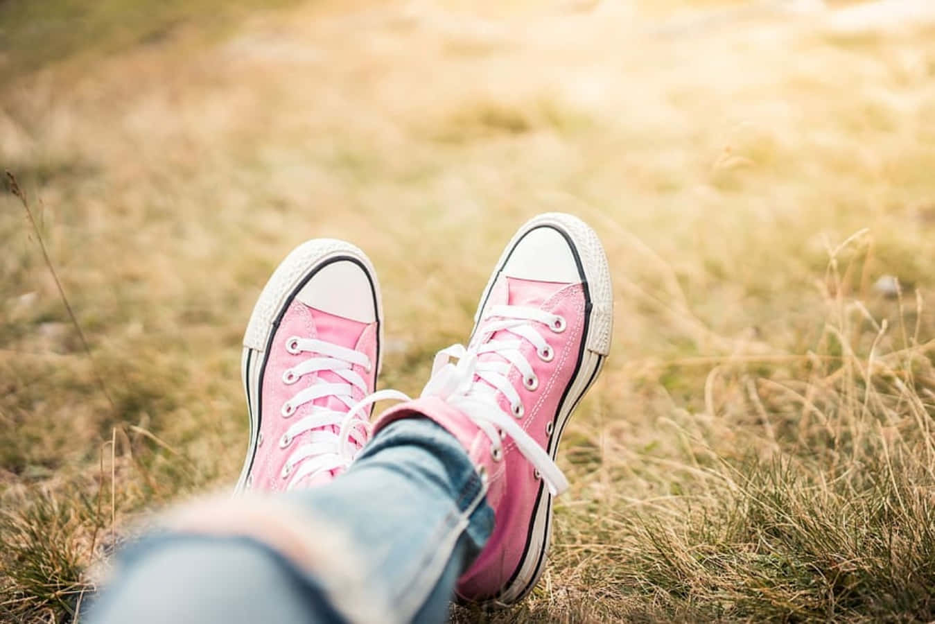 Pink Shoes 桌布 壁紙 — 粉紅色運動鞋搭配白色鞋帶擱在金黃色的草地上，雙腿在陽光明媚的田野中放鬆地晃動。