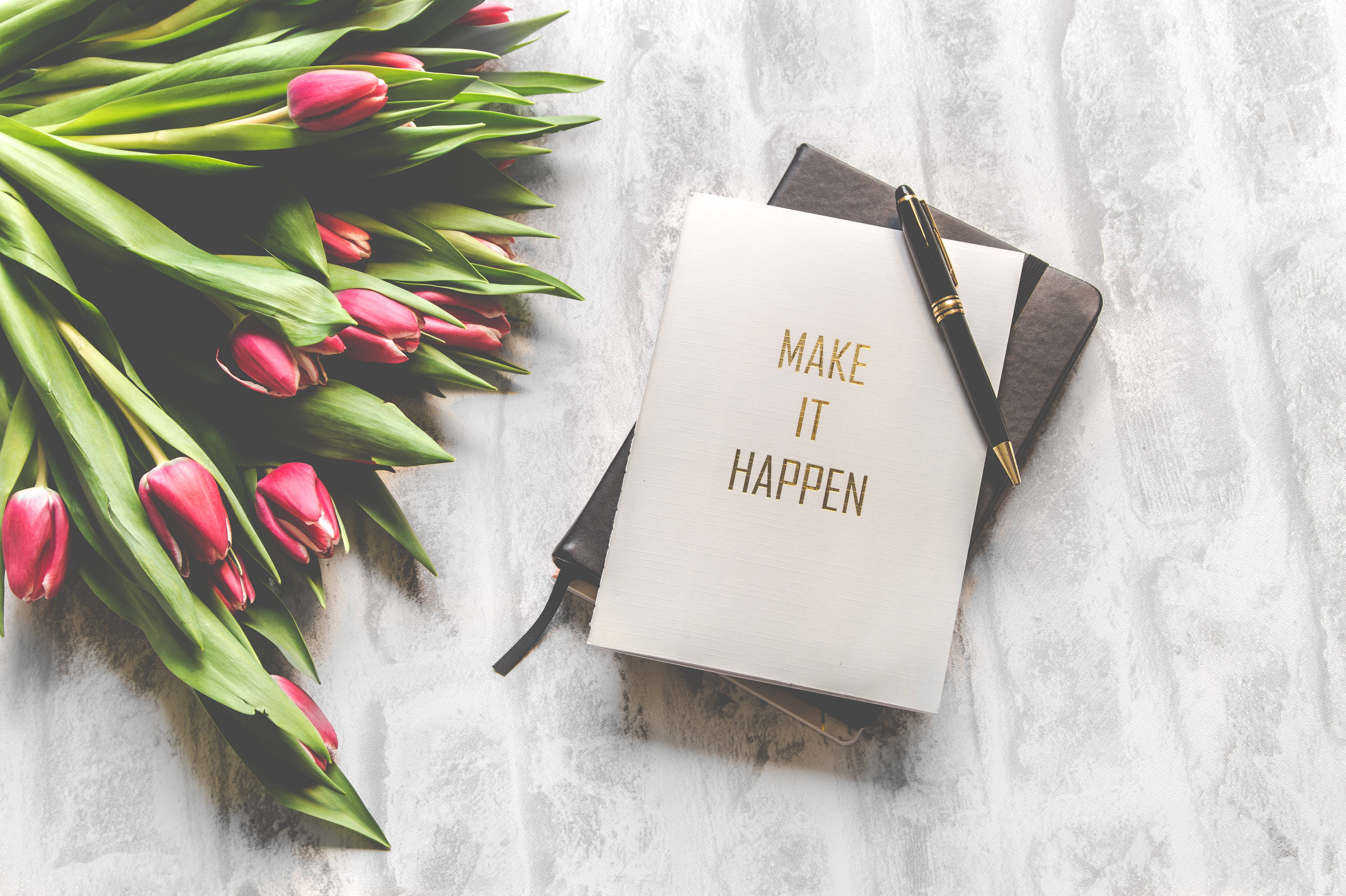 鬱金香 桌布 壁紙 — 粉紅色鬱金香和一本寫有「MAKE IT HAPPEN」的筆記本，放在有紋理的白色表面上，呈現寧靜的平鋪佈局。