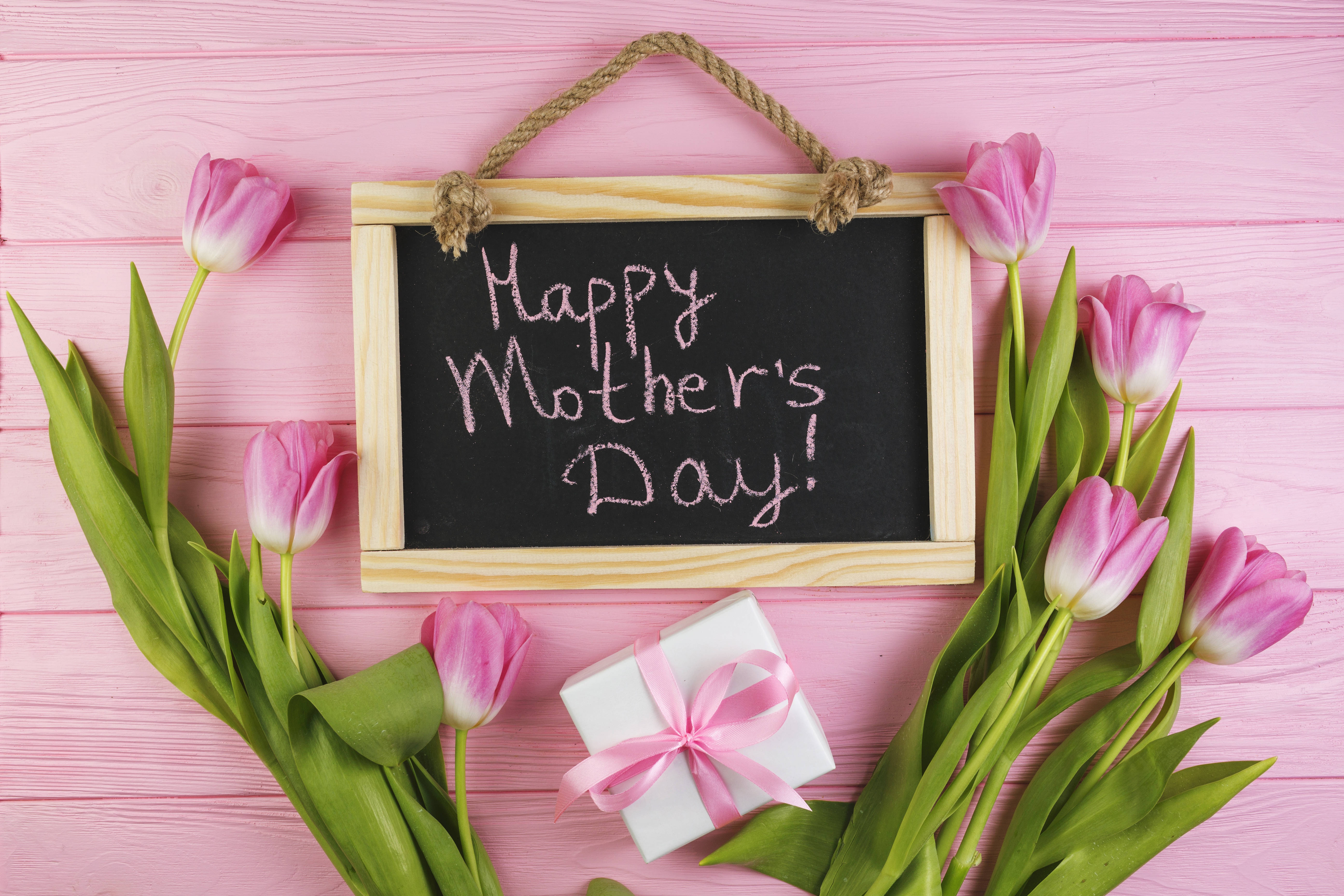 Happy Mothers Day 桌布 壁紙 — 粉紅色鬱金香和禮物盒圍繞著一塊寫有「Happy Mother's Day!」的黑板，放在粉紅色木質表面上。