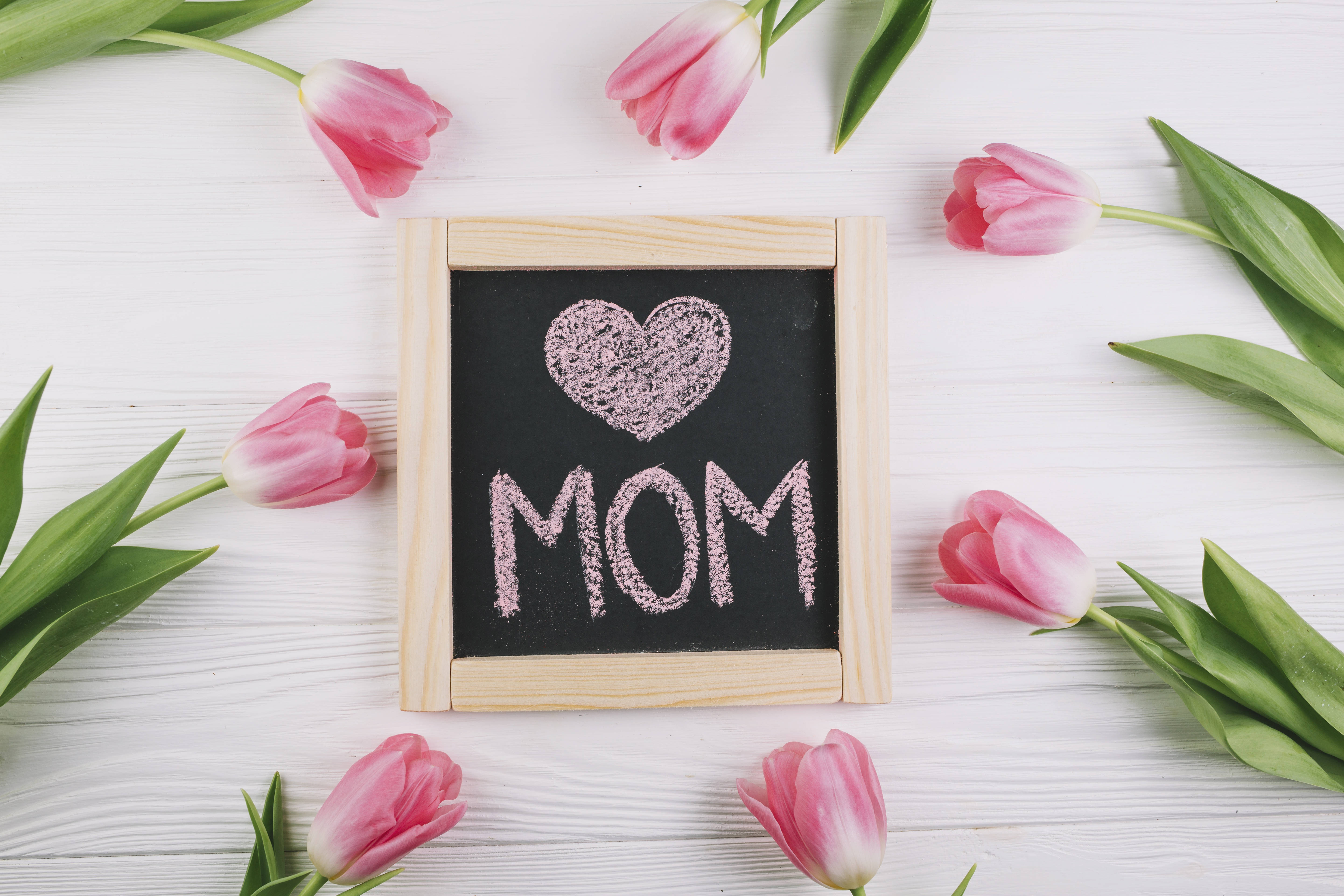 Happy Mothers Day 桌布 壁紙 — 粉紅色鬱金香圍繞著一個木框黑板，上面用粉紅色粉筆在白色木質表面上寫著一個心形和「MOM」。