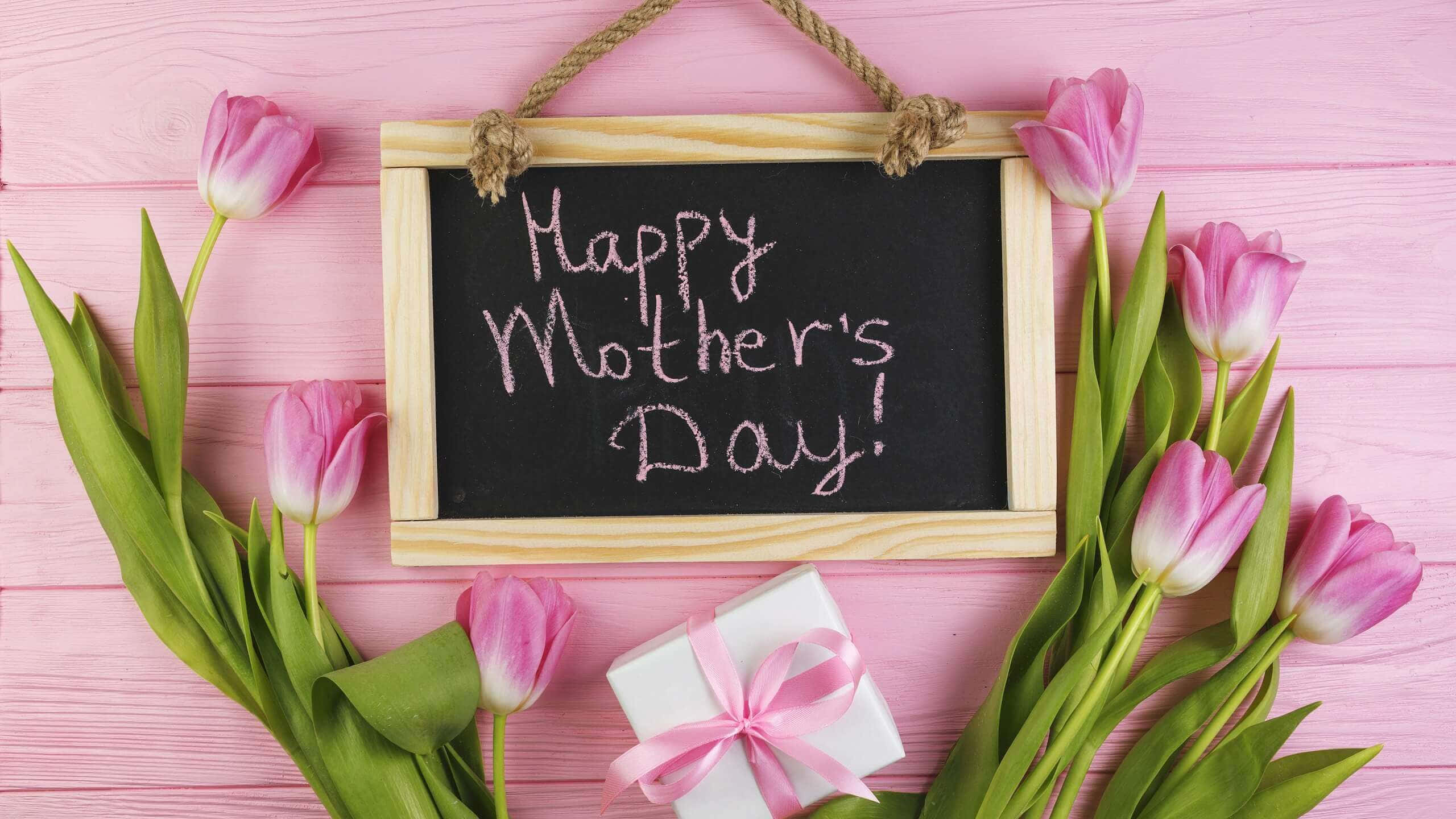 happy mother's day 桌布 壁紙 — 粉紅色鬱金香圍繞著一塊寫有「Happy Mother's Day」的黑板標誌，背景是粉紅色木質，並附有一個禮物盒。