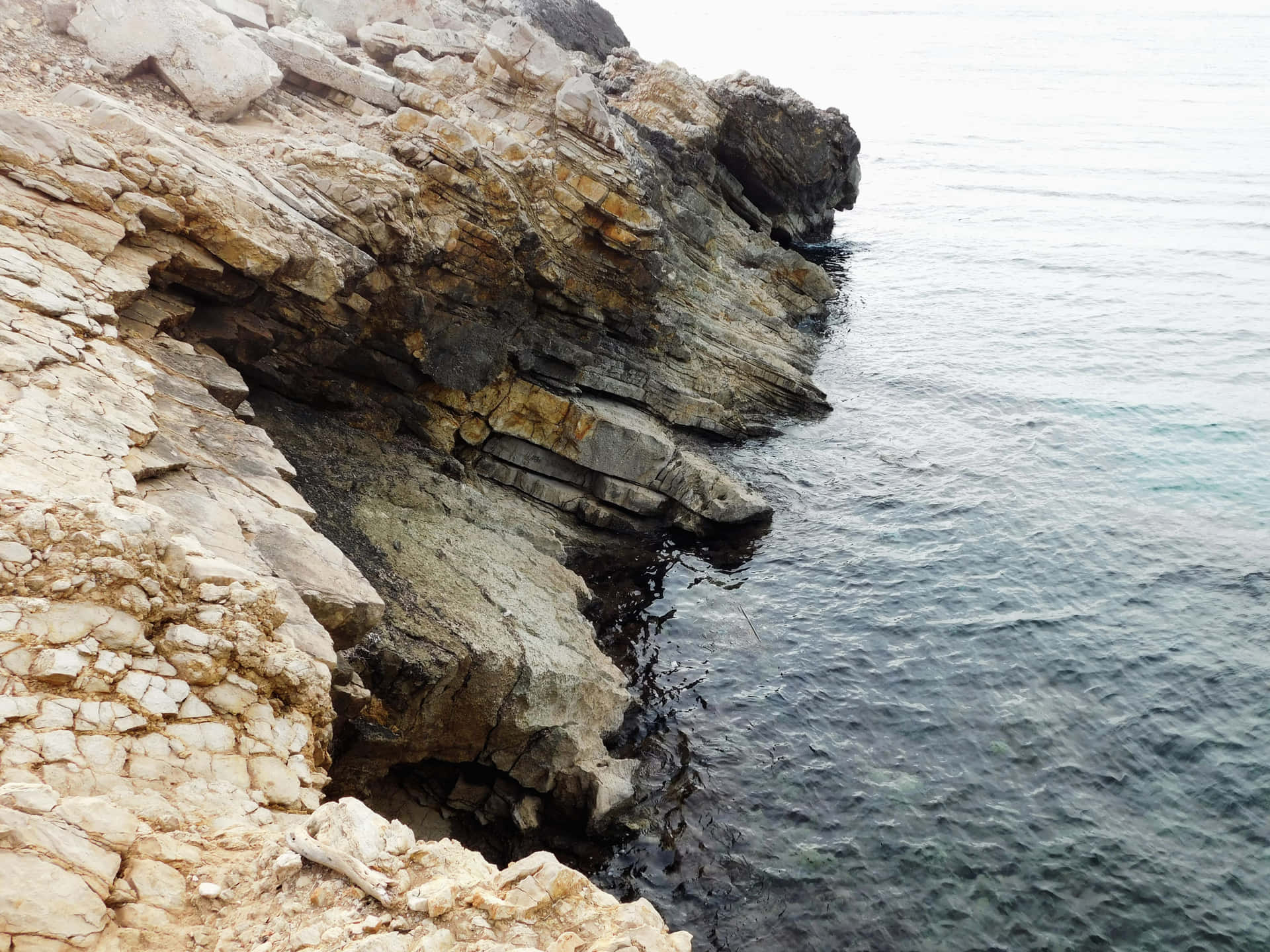 Sea with rocks 壁紙 — 粗糙層狀的米灰色和赭色石崖在柔和陰天下與平靜的碧綠海水相遇。