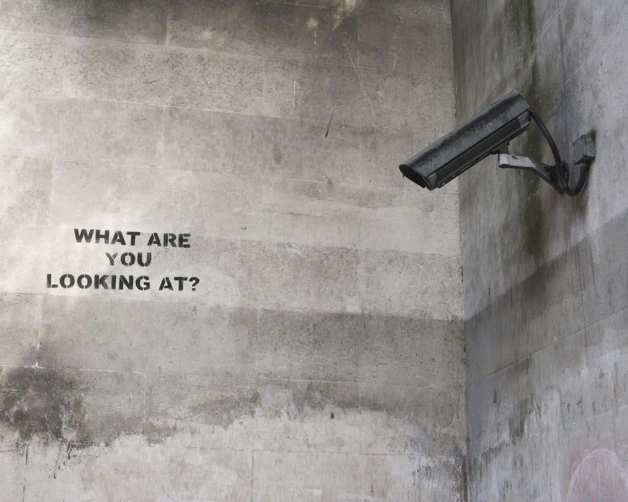 Banksy 桌布 壁紙 — 粗糙的混凝土牆上，在安裝的監控攝影機正下方，有粗體塗鴉文字問『你在看什麼？』