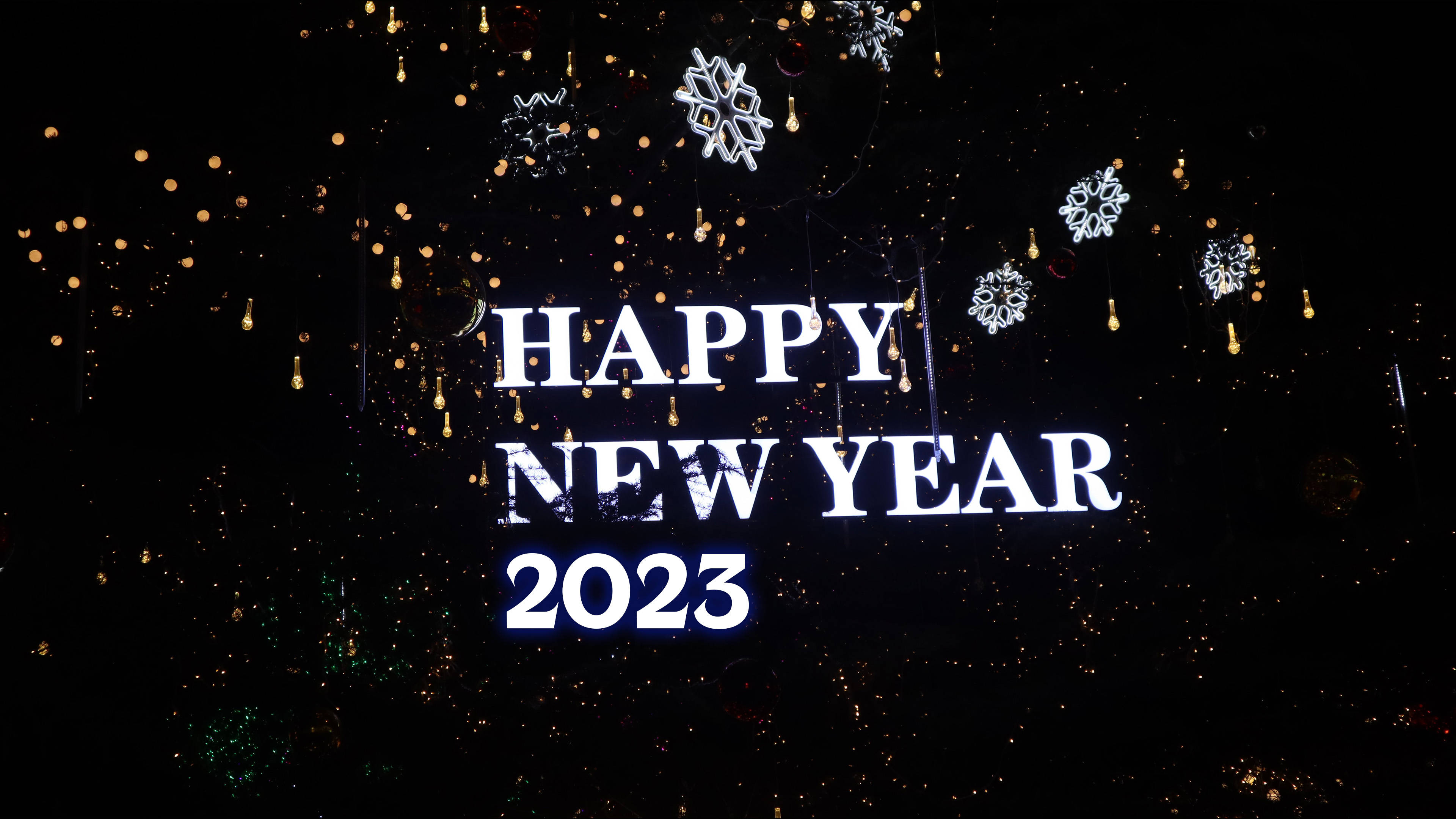 新年快樂 桌布 壁紙 — 粗體白色 'HAPPY NEW YEAR 2023' 文字在黑暗背景上發光，背景點綴著閃爍的金色燈光和懸掛的雪花裝飾。