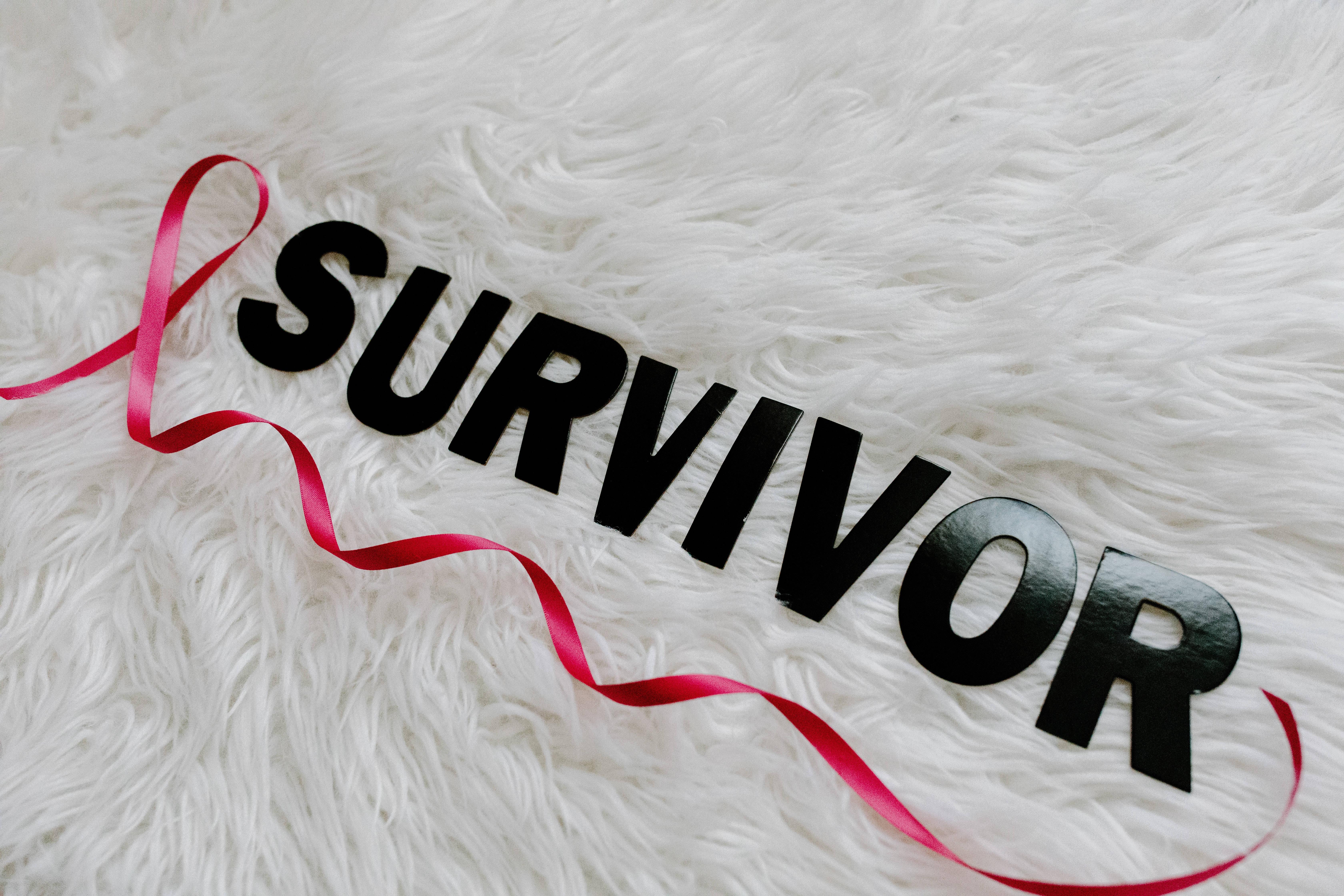 白色 桌布 壁紙 — 粗體黑色 'SURVIVOR' 字母和一條紅色絲帶放在柔軟的白色絨毛表面上。