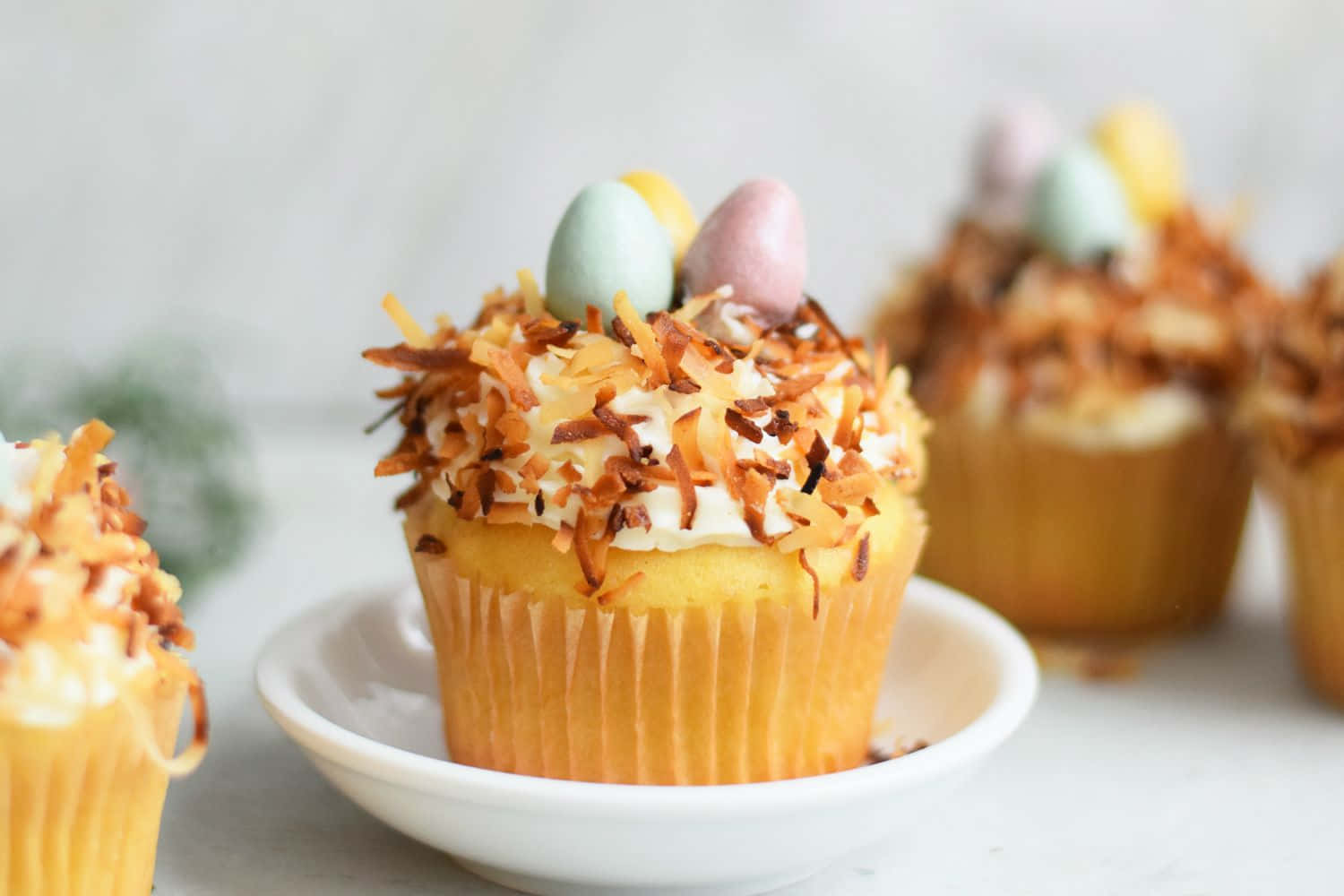 Cupcake 桌布 壁紙 — 精緻的杯子蛋糕，上面撒有烤椰子片和粉彩糖果蛋，放在白色盤子上，背景柔和模糊。