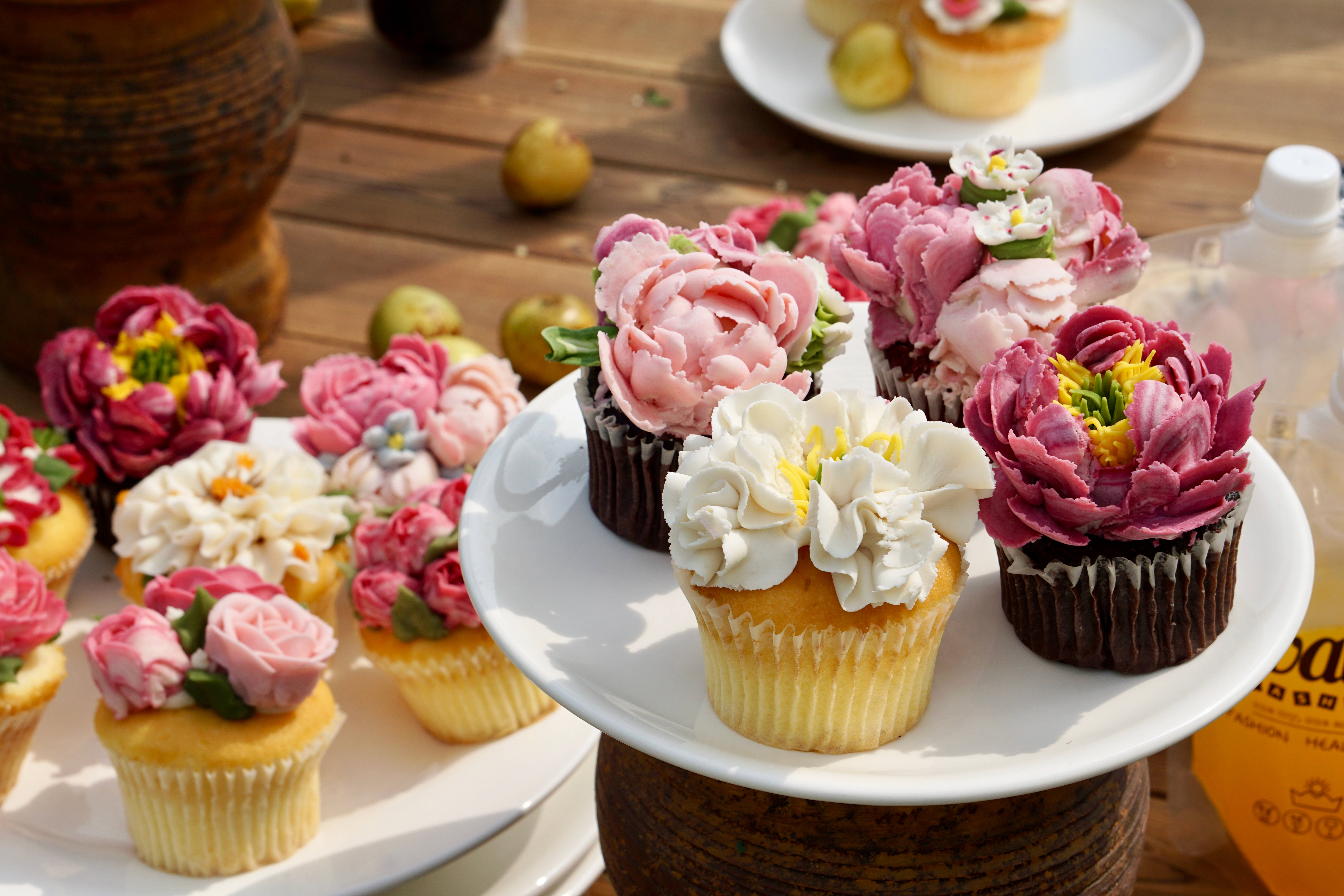 Cupcake 桌布 壁紙 — 精緻的杯子蛋糕上綴有粉紅、白色和紫色的精細糖花，在明亮的陽光下綻放在木桌上。