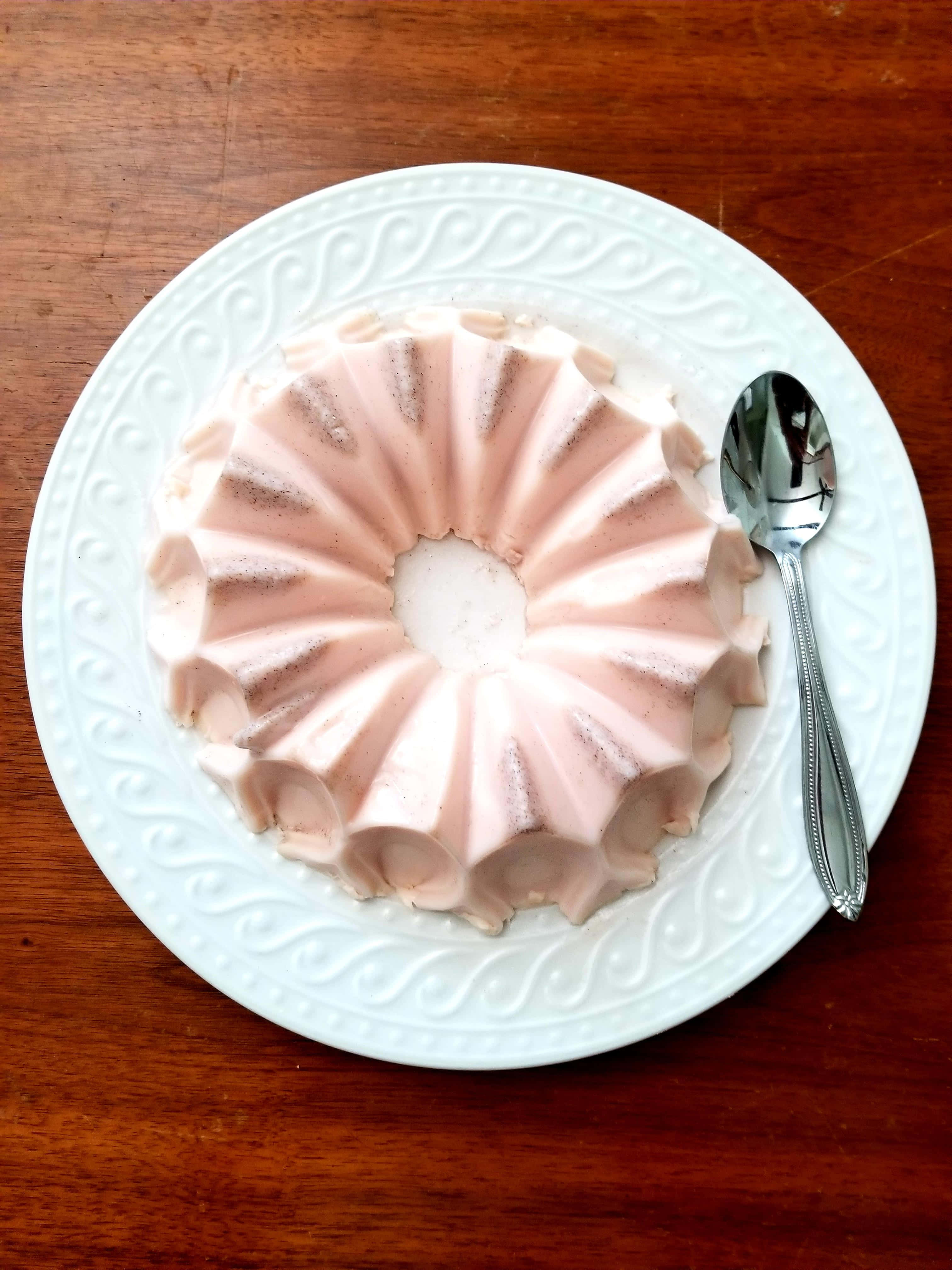 Blancmange 桌布 壁紙 — 精緻的粉紅色蛋白霜甜點撒上糖粉，放在裝飾性的白色盤子上，旁邊有銀湯匙，置於木桌上。