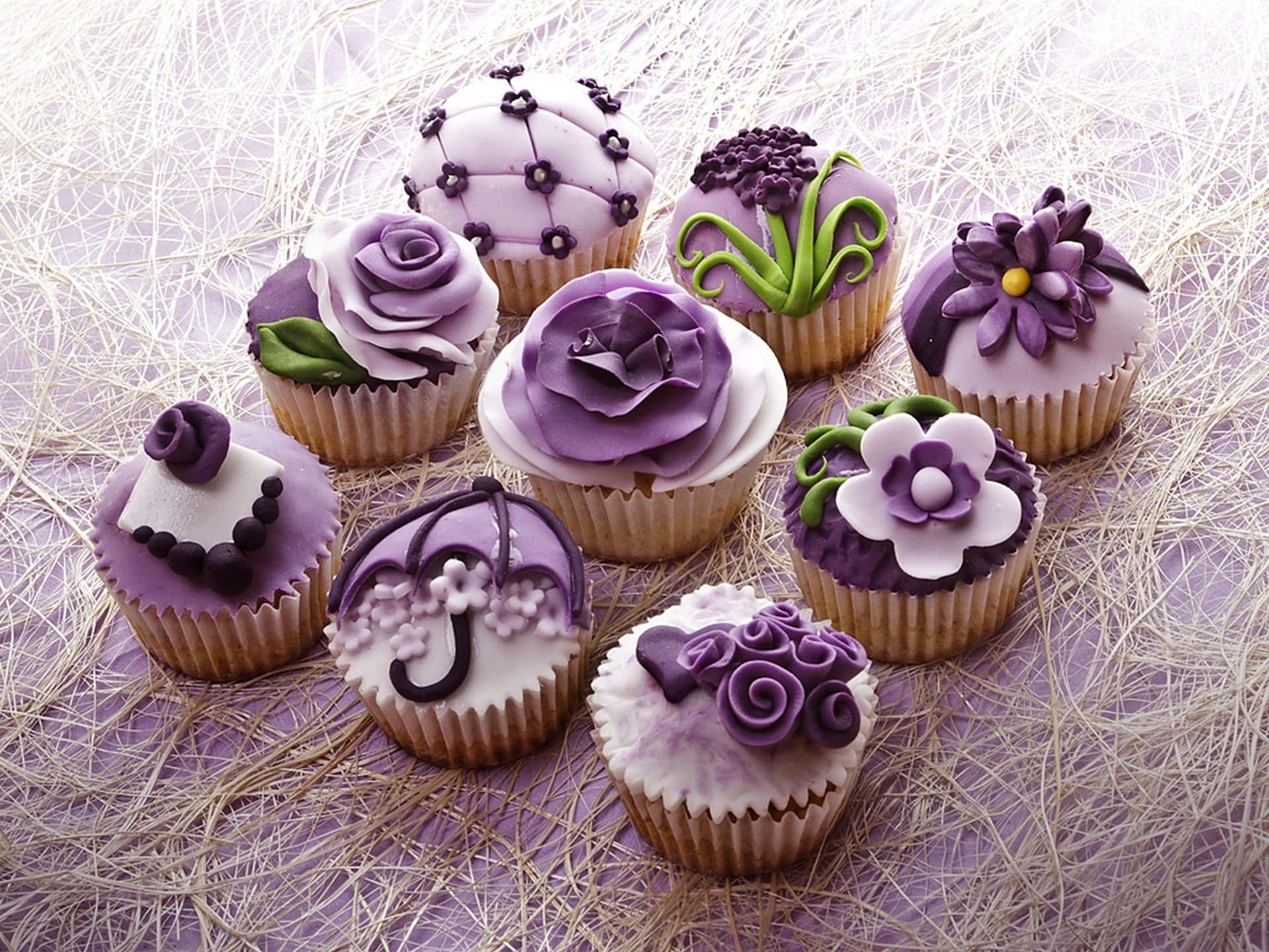 Spring Desserts 桌布 壁紙 — 精緻的紫色紙杯蛋糕，飾以糖花和蕾絲細節，躺在稻草床上，襯以柔和的薰衣草背景。
