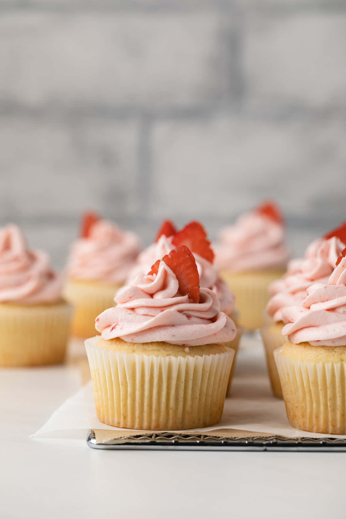 Cupcake 桌布 壁紙 — 精緻的草莓杯子蛋糕，配上粉紅色糖霜和新鮮草莓片，放在白色表面上，背景是柔和的灰色磚牆。