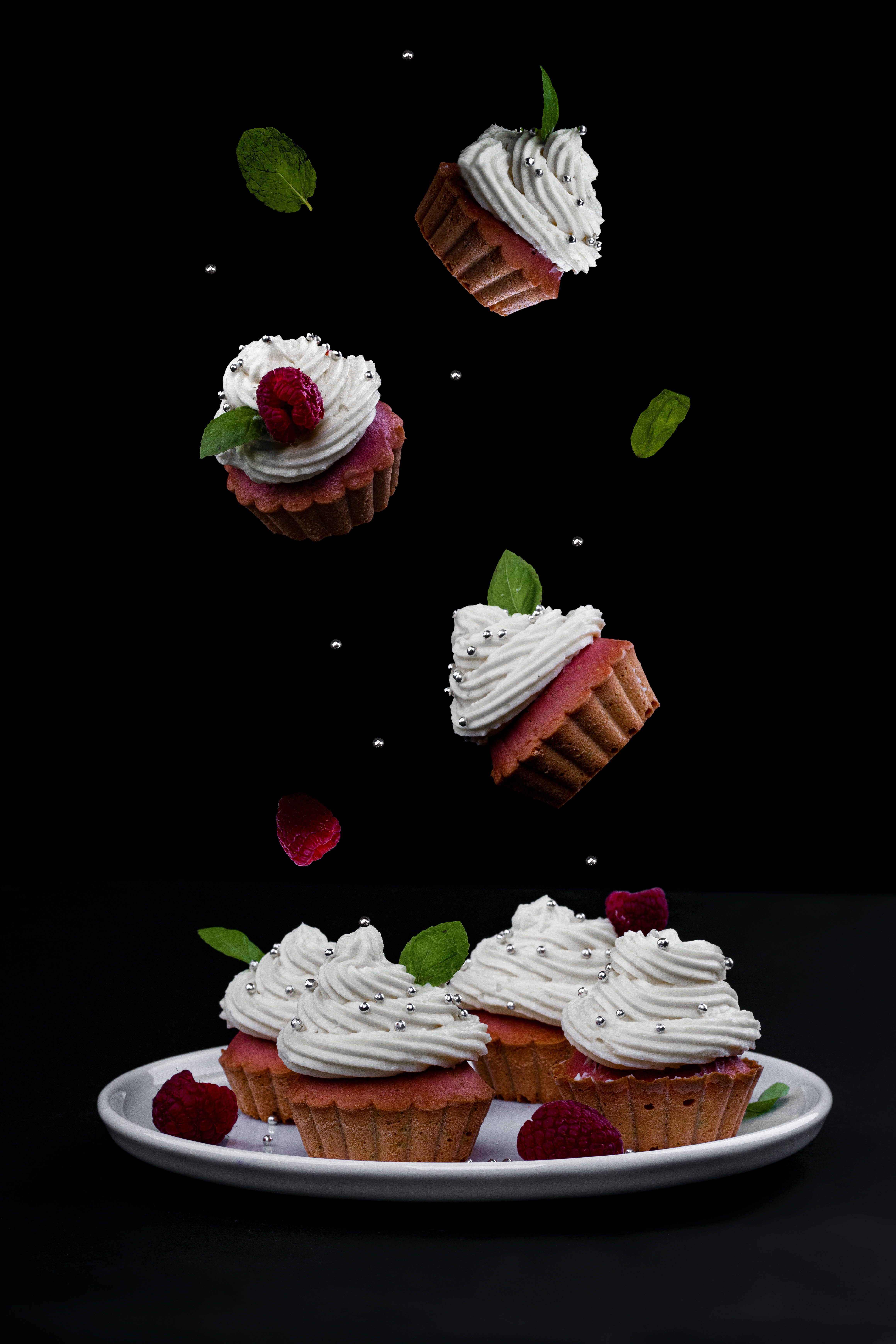 Cupcake 桌布 壁紙 — 精緻的覆盆子杯子蛋糕，配上白色糖霜和薄荷葉，懸浮在空中，位於白色盤子上方，背景為黑色。