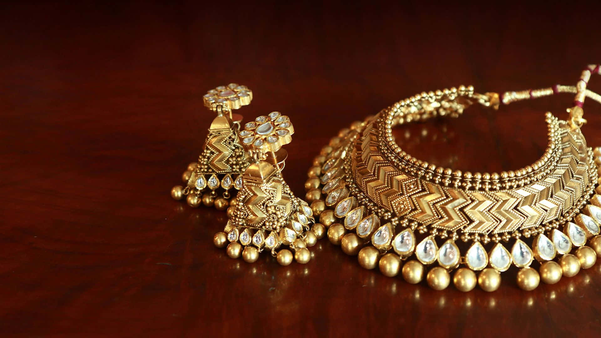 Earrings 桌布 壁紙 — 精緻的金色珠寶套裝，帶有複雜圖案和垂墜寶石，放置在濃郁的桃花心木表面上。