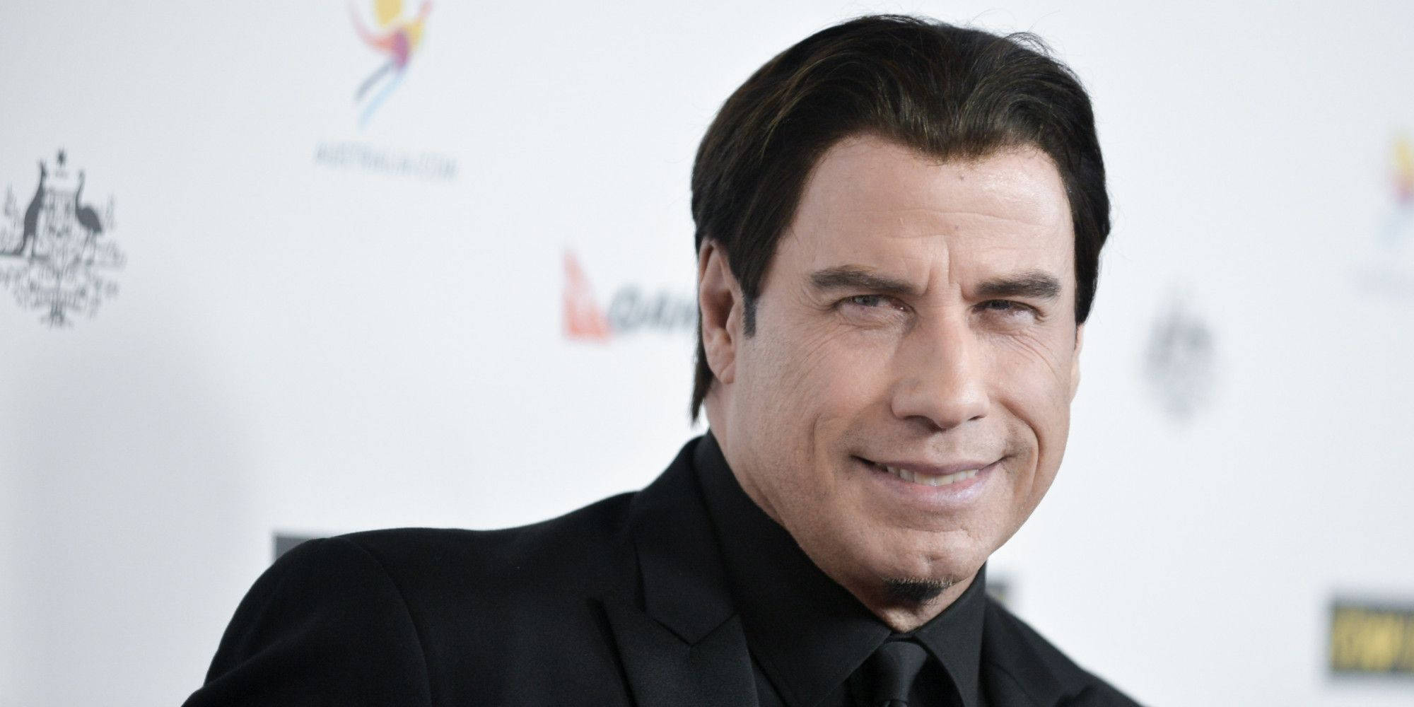 Celebrity 桌布 壁紙 — 約翰屈伏塔身穿黑色西裝，在帶有標誌的白色活動背景前自信微笑。