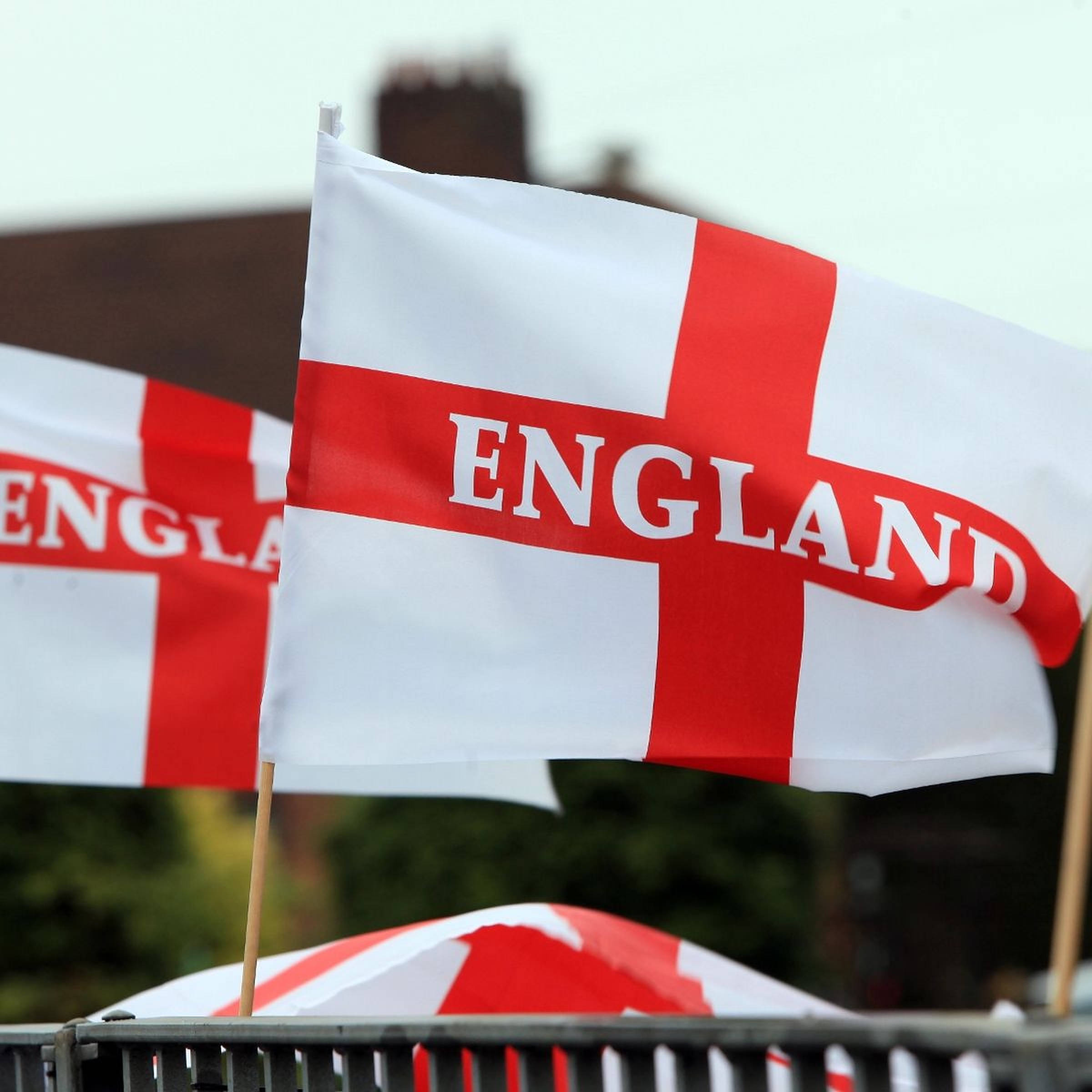 England Flag 桌布 壁紙 — 紅白英格蘭旗幟隨風飄揚，粗體字樣映襯模糊的戶外背景。