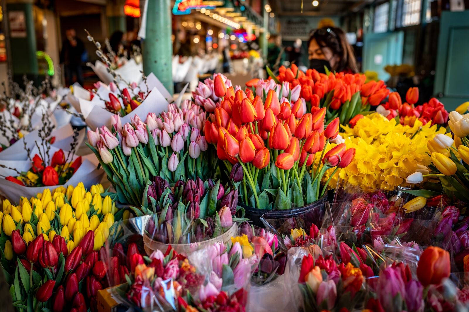 Pike Place Market 桌布 壁紙 — 紅色、粉紅色、黃色和紫色的鮮豔鬱金香花束，充滿了繁忙的室內花卉市場攤位。